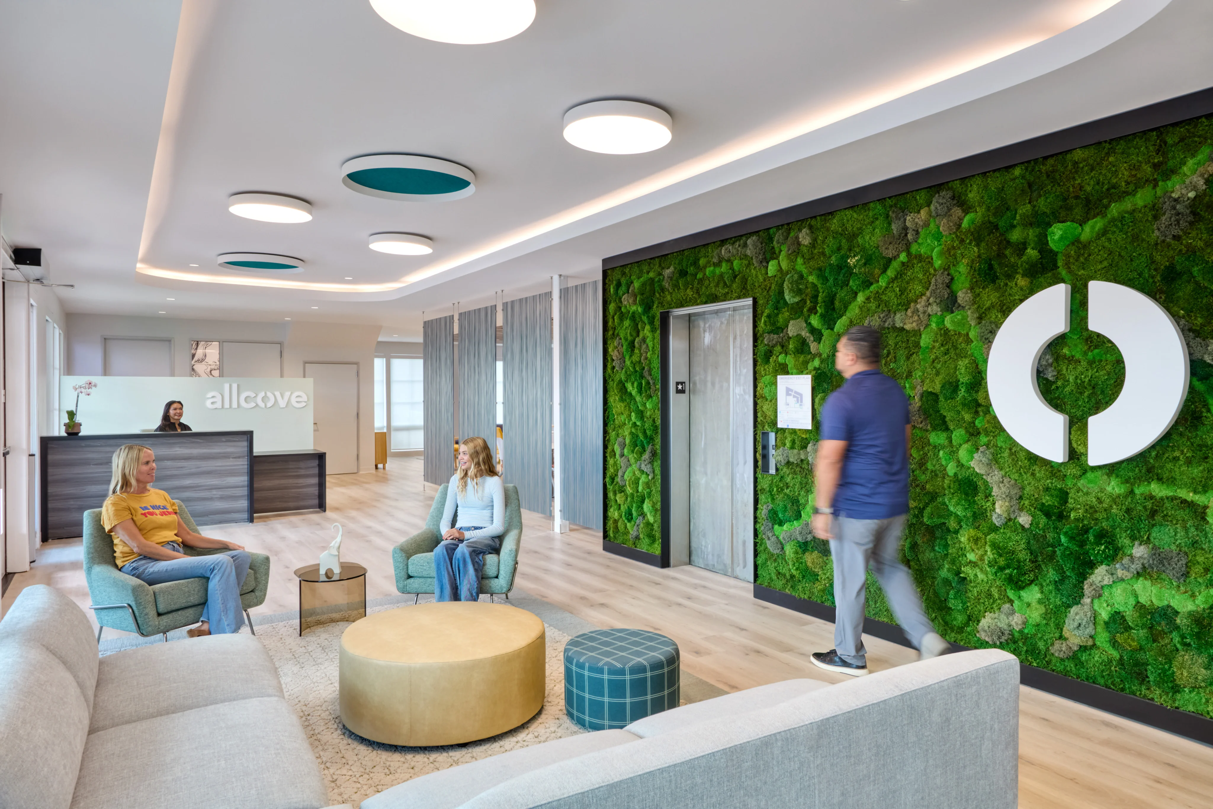 Modern office lobby with a green moss wall, sleek ceiling lights, and a reception desk. Two people sit on chairs, another walks past, creating a calm and professional atmosphere.