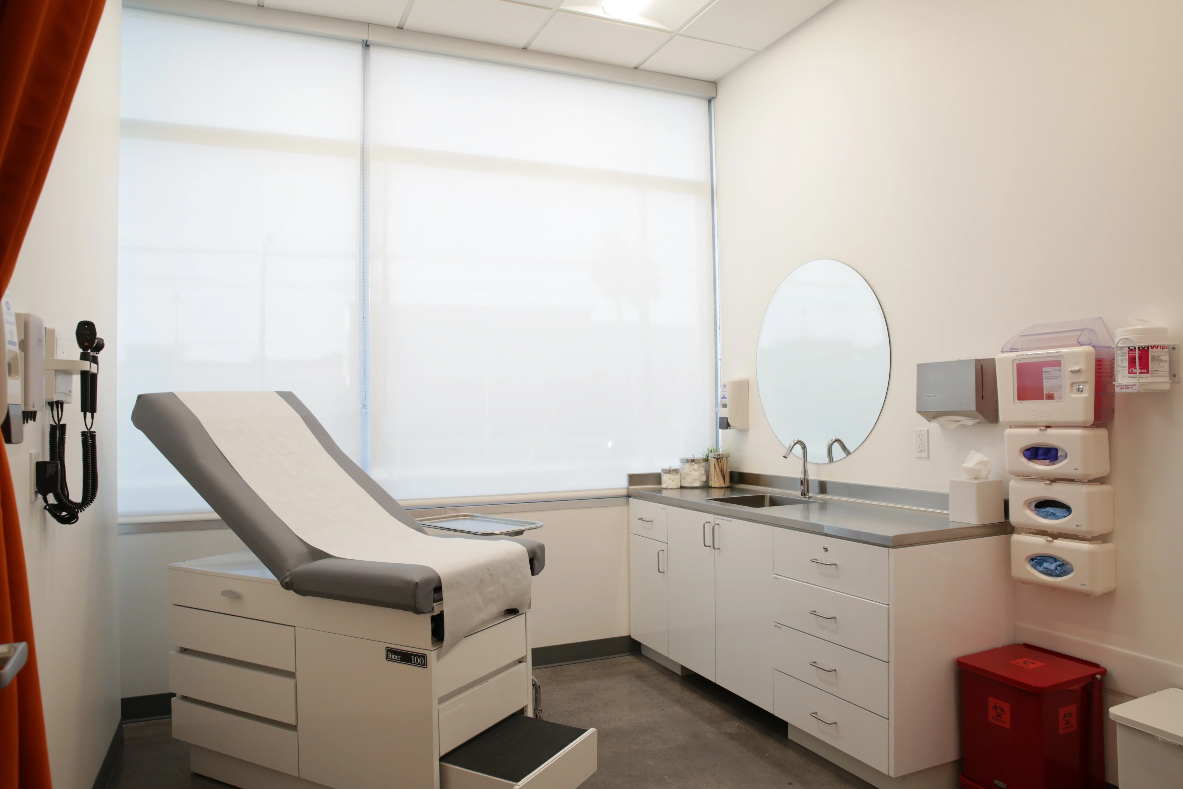 A clean, bright medical exam room with a patient table, white cabinets, a round mirror, and medical supplies. An organized, professional setting.