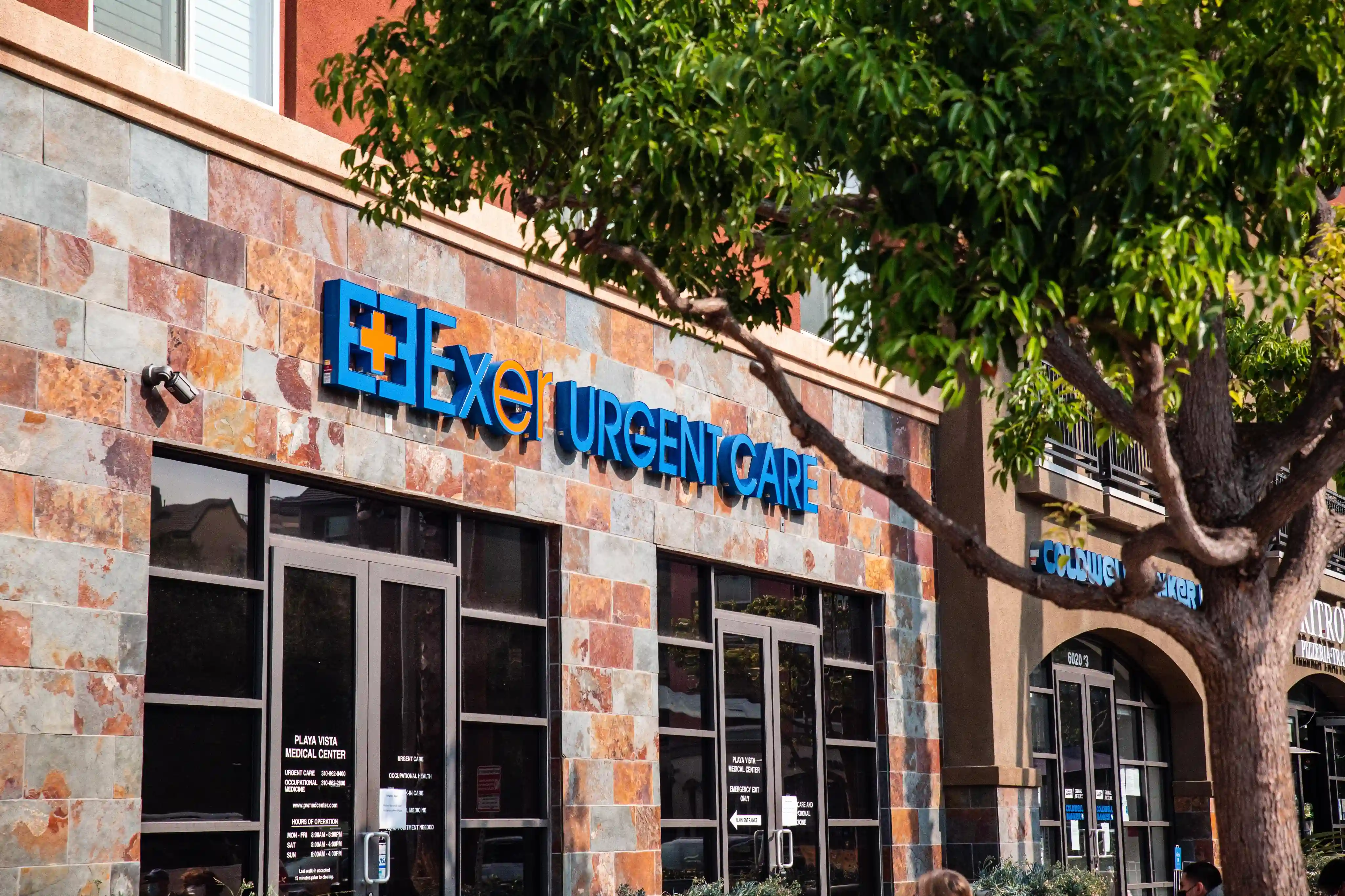 Exterior of an urgent care clinic with a stone facade and blue signage. Large tree partially shades the entrance, creating a calm, inviting atmosphere.