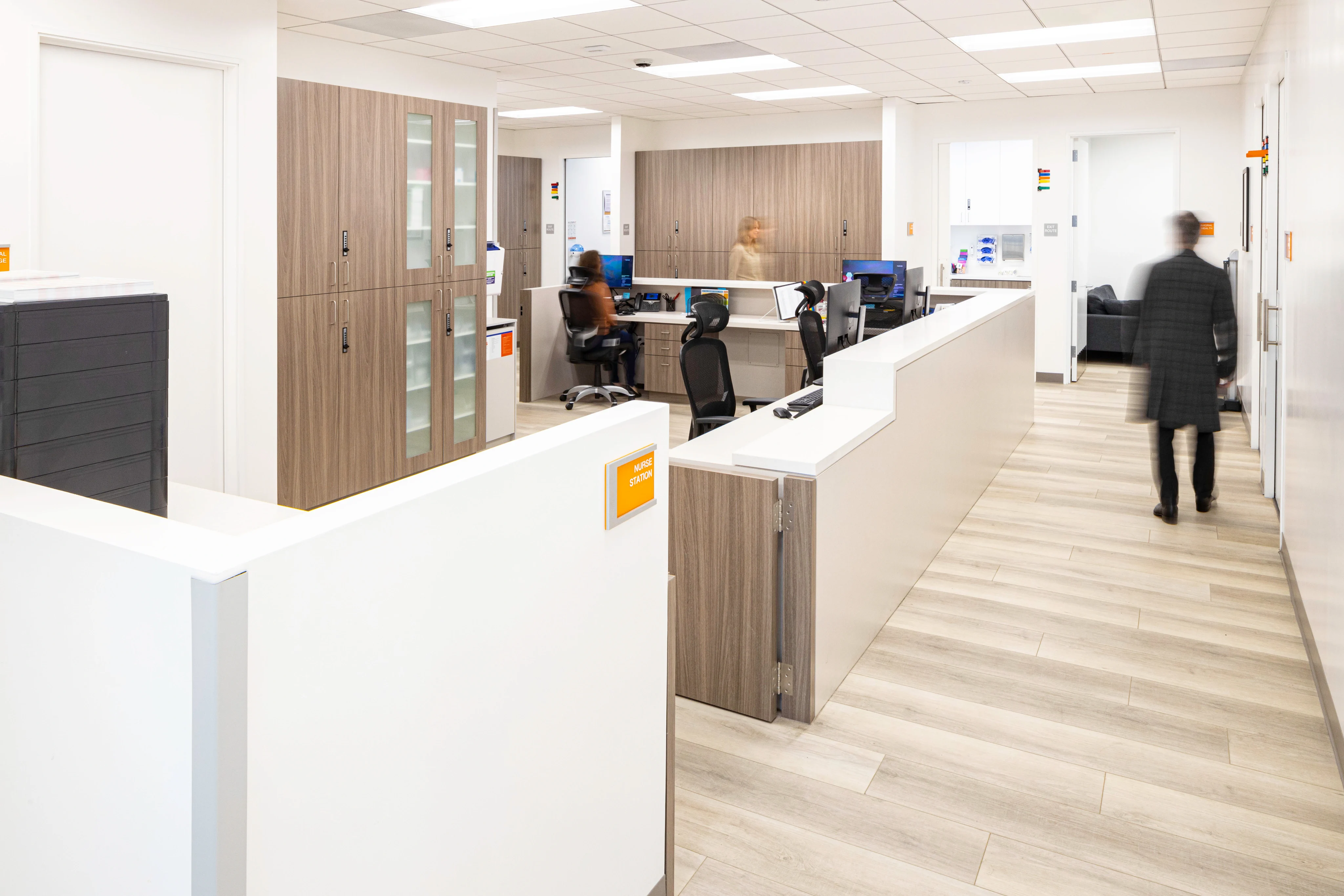 Modern office space with white walls and wood accents, featuring workstations, computers, and a few blurred figures walking. Bright, organized atmosphere.