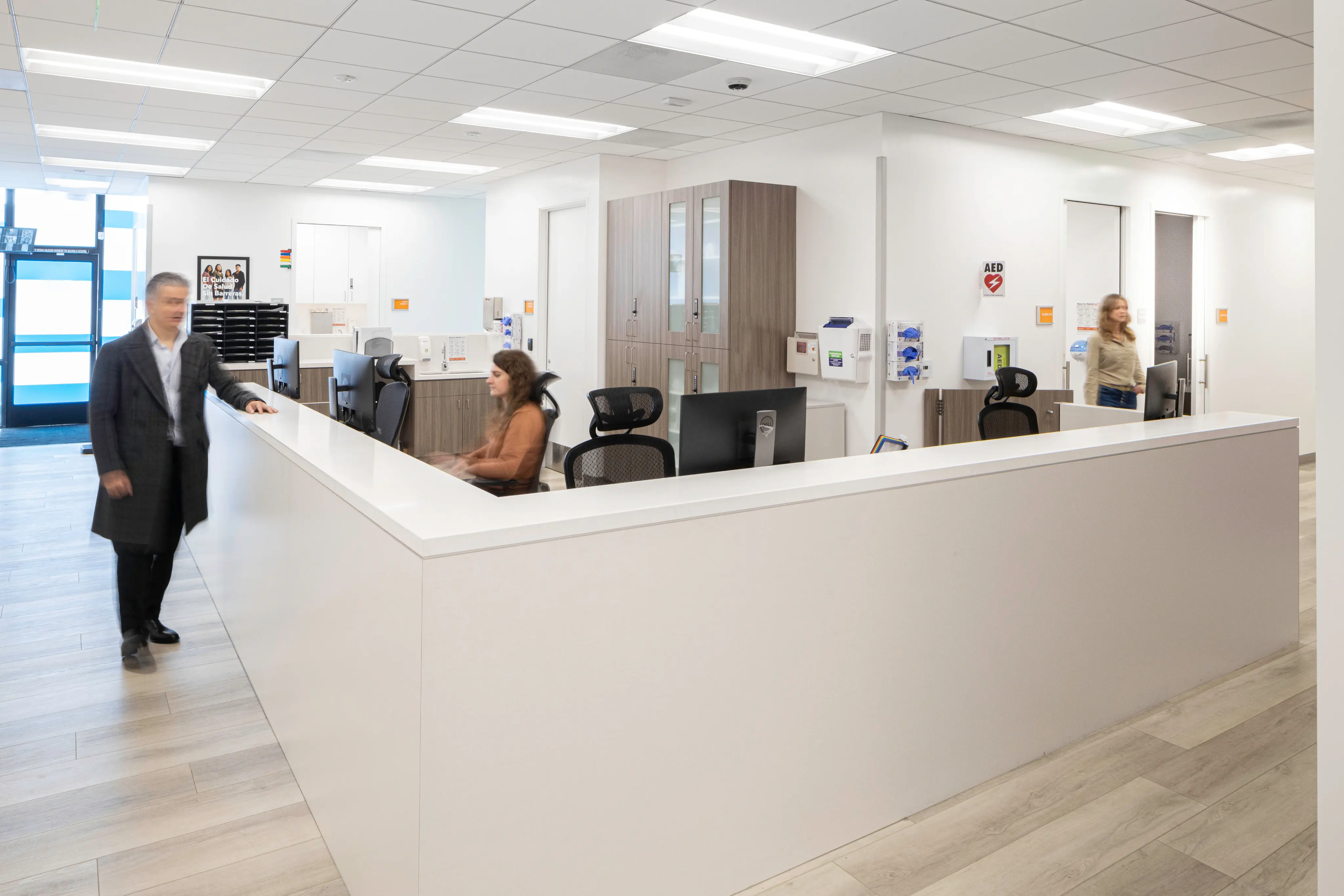 Modern office reception with a white desk and wood cabinets, featuring two blurred figures walking and a seated person working. Bright, professional ambiance.