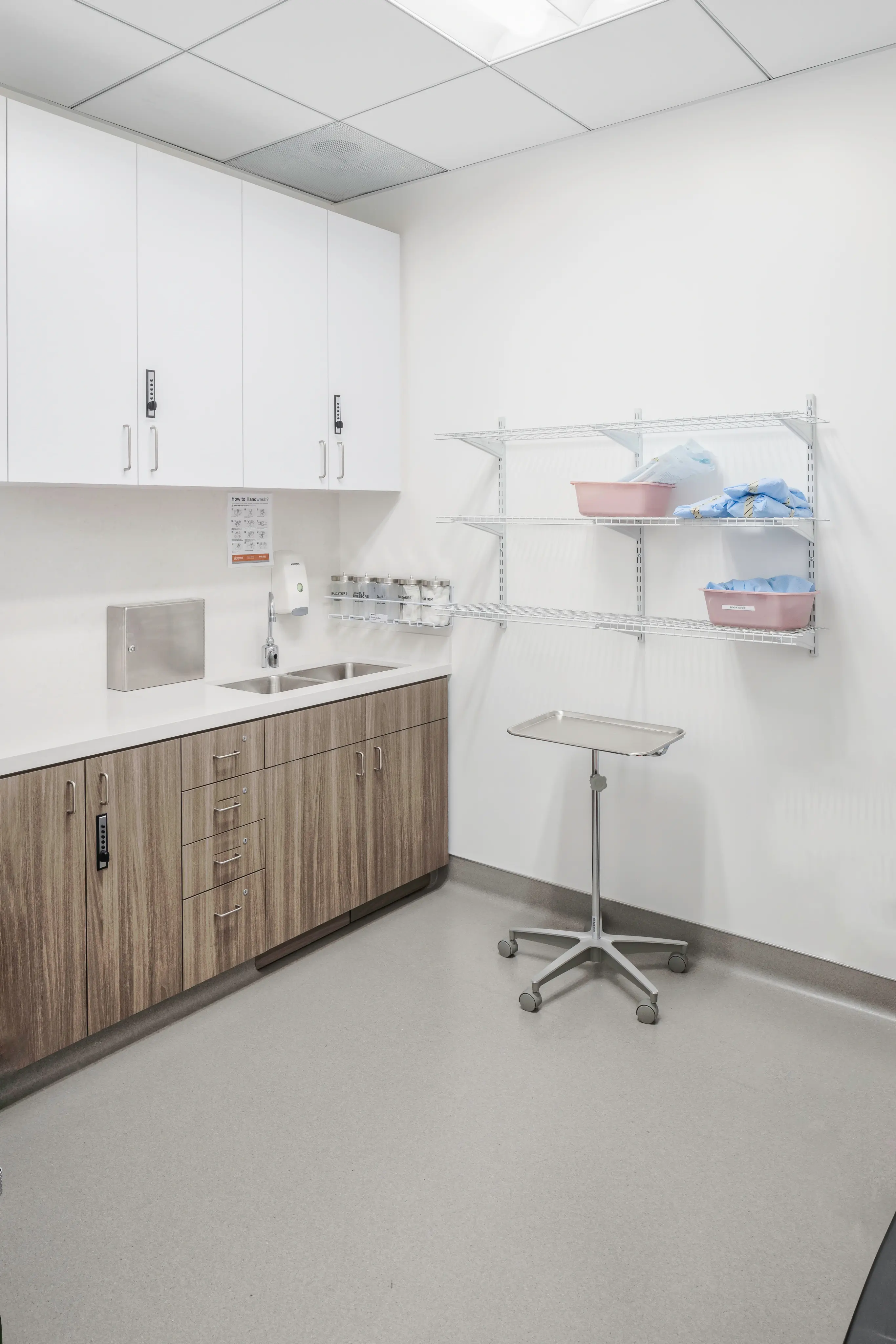 A sterile medical room with white cabinets, a steel sink, and wooden lower cabinetry. Shelves hold medical supplies, while an adjustable tray table stands nearby.