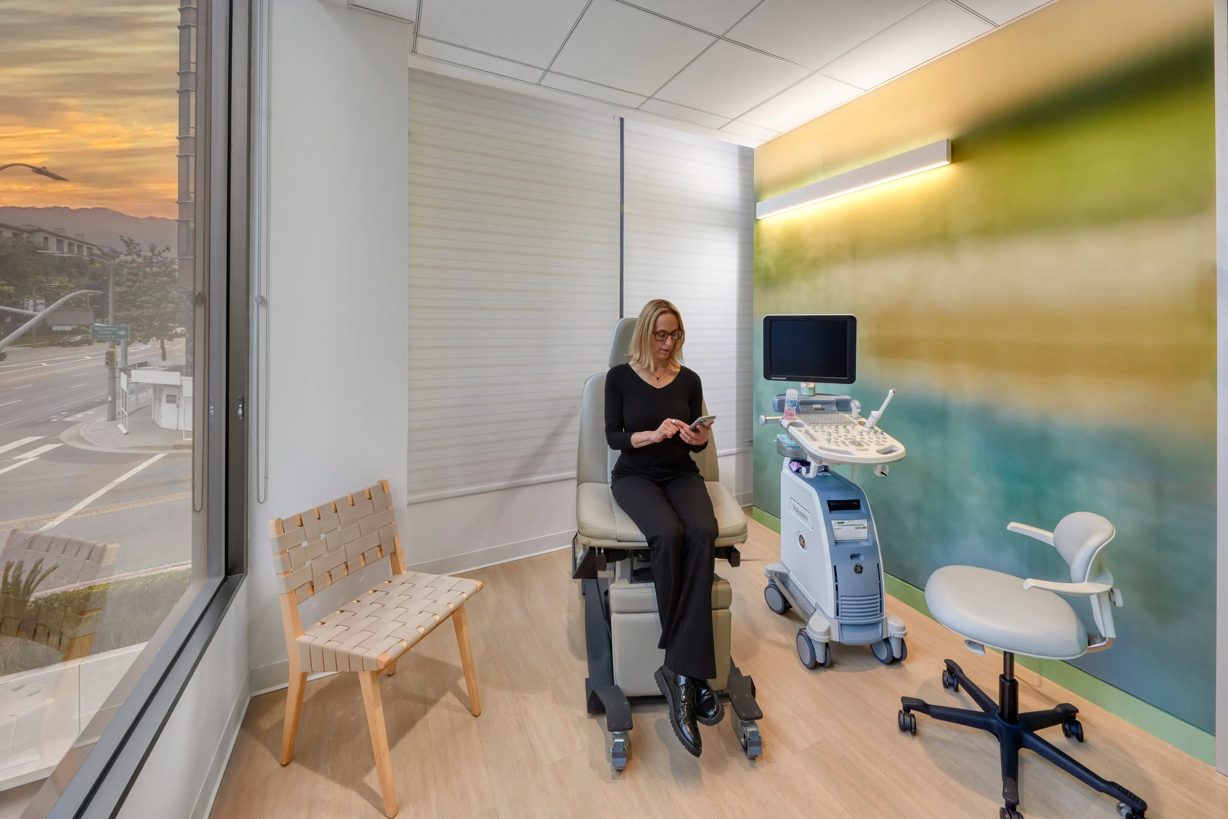 A woman sits on an exam chair in a modern medical room, using her phone. There's an ultrasound machine nearby and a sunset visible through the window.