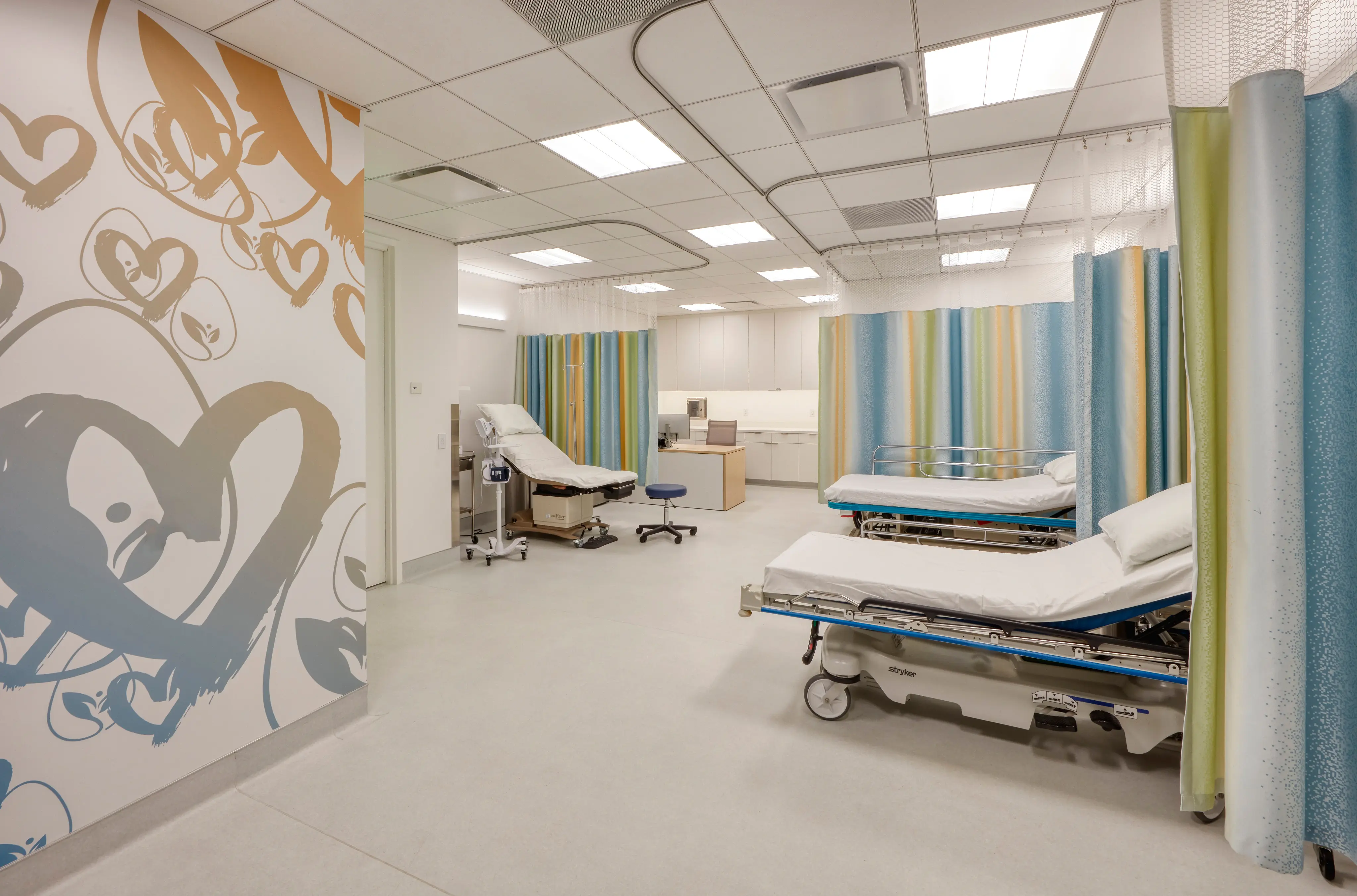 A bright, modern hospital room with two patient beds, blue and green privacy curtains, heart design on feature wall, and ambient fluorescent lighting.