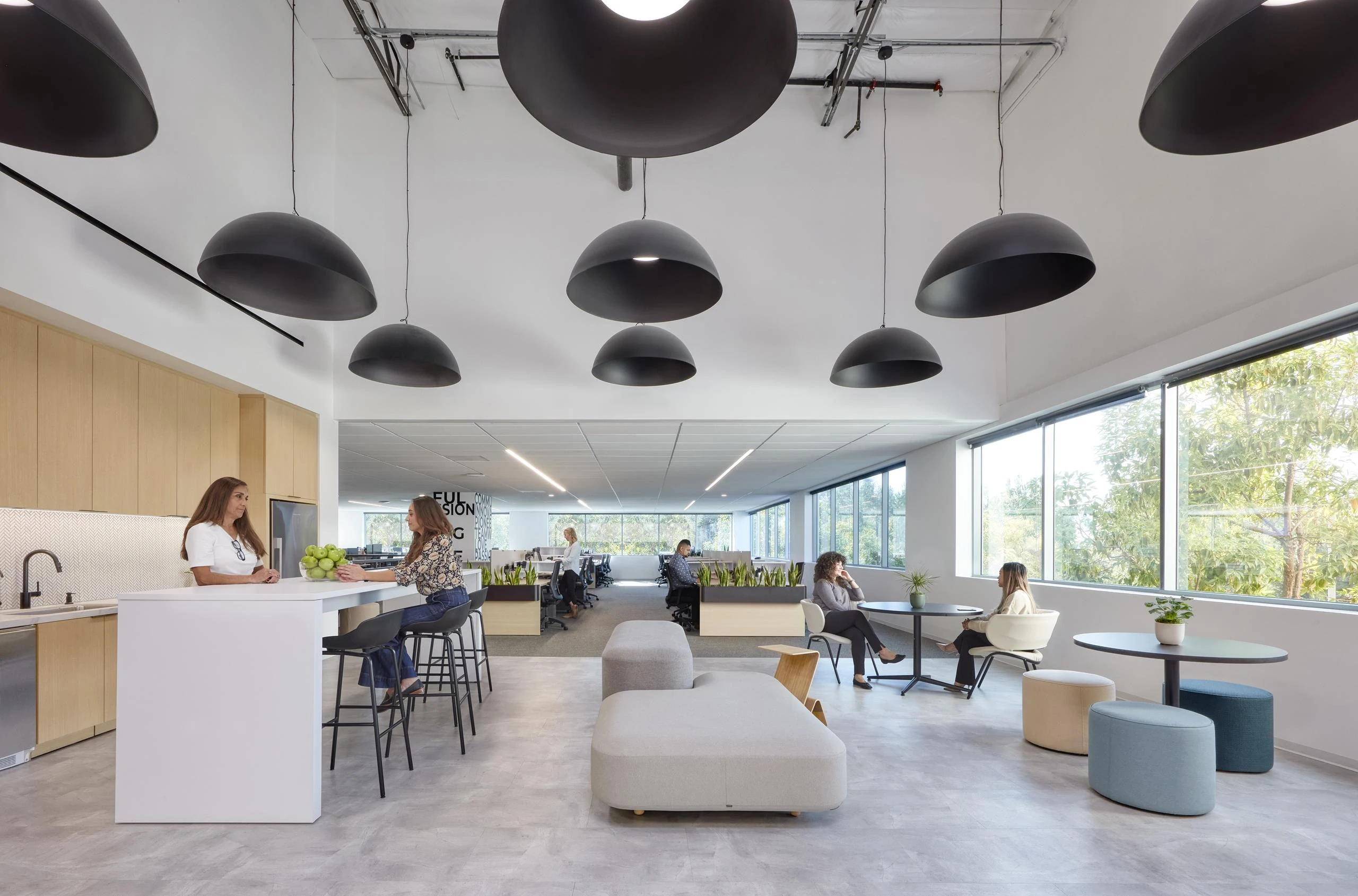 Modern open-plan office with sleek black pendant lights, light wood tones, and large windows. People sit casually on stylish furniture, creating a relaxed atmosphere.