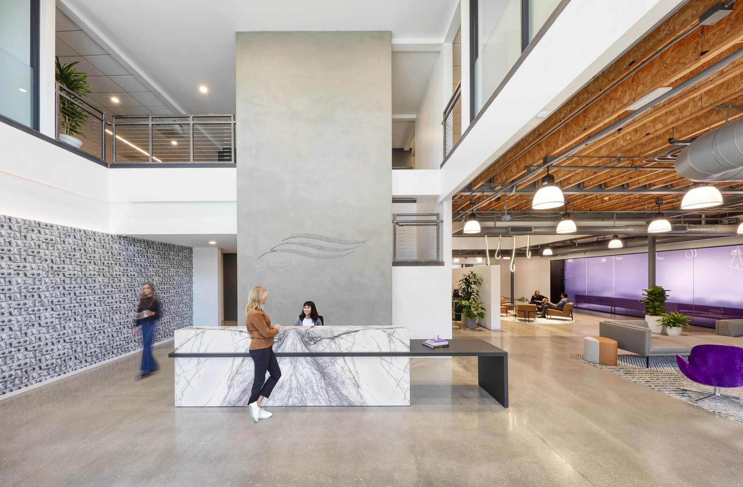 Modern office lobby featuring a sleek marble reception desk, two people conversing, open seating area with purple accents, and high ceilings.
