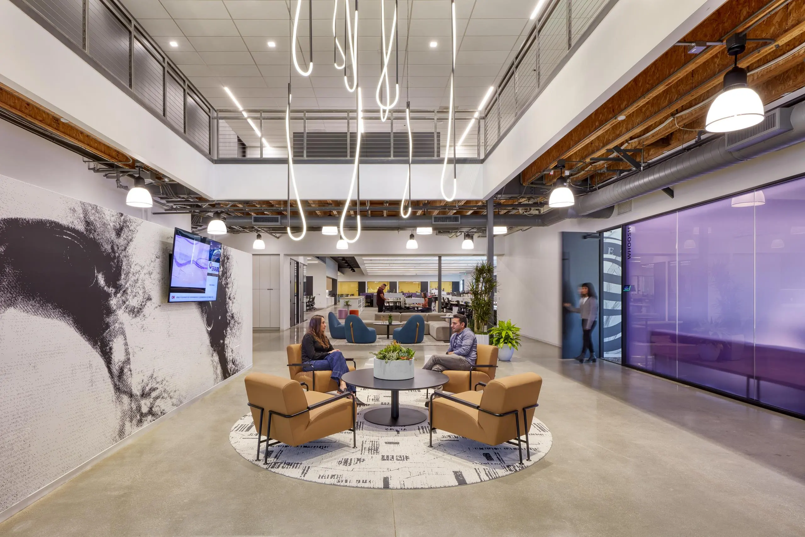 Modern office lobby with two people conversing on tan chairs around a circular table. Artistic lighting hangs from ceiling; a purple glass wall is visible.
