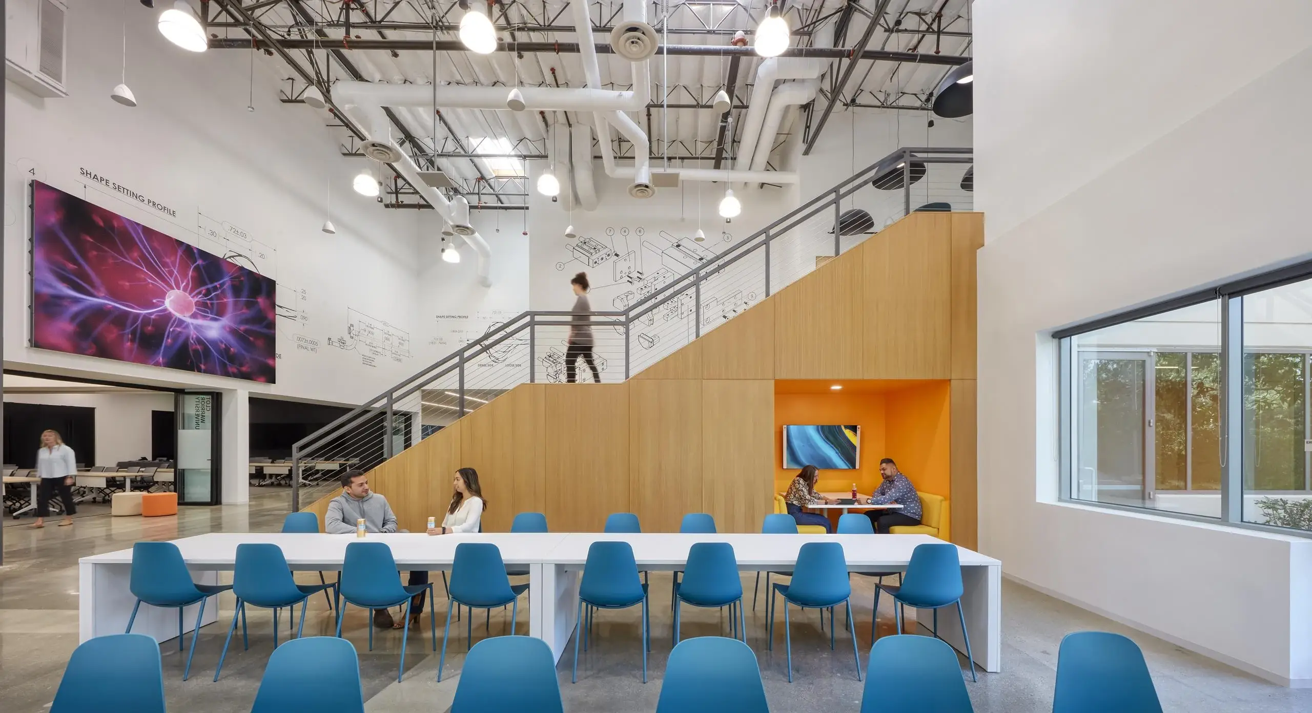 Modern office space with a high ceiling features a long white table with blue chairs, a large digital screen, staircase, and people engaging in conversation.