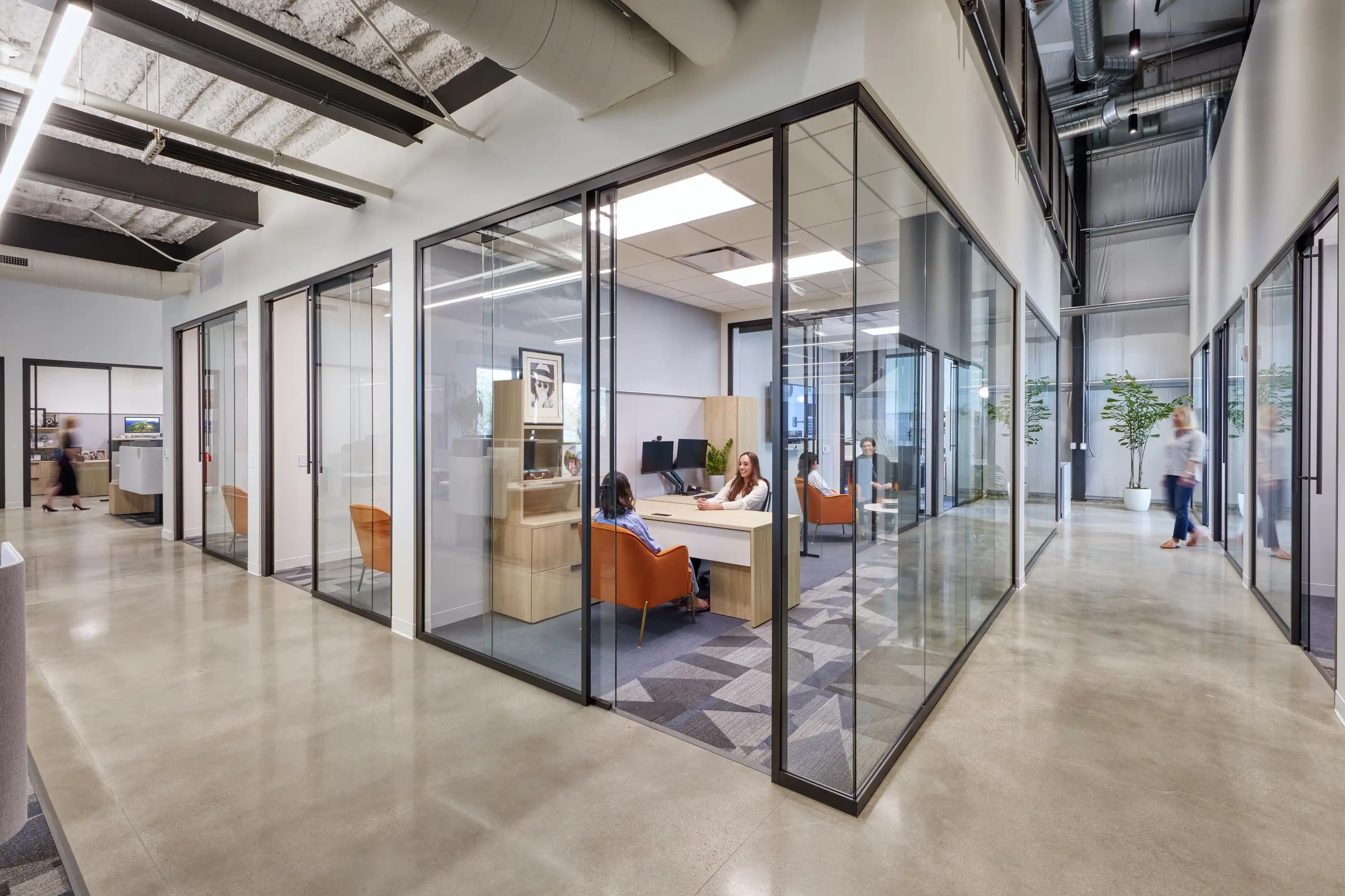 Modern office space with glass-walled meeting rooms. Two people are seated, discussing at a desk. The area is bright, open, with plants adding a touch of nature.