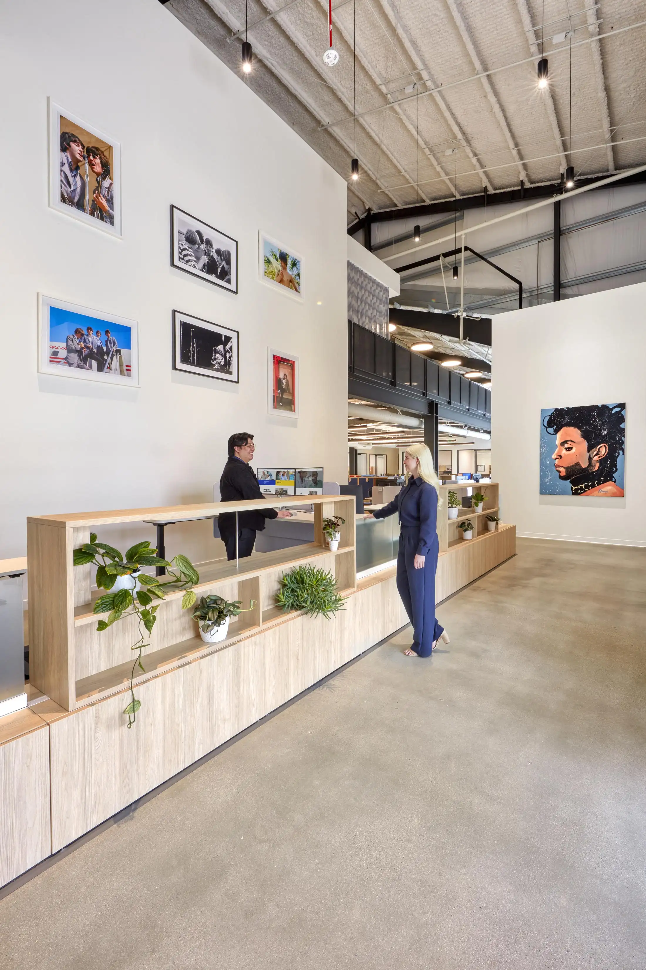 A modern office lobby with tall ceilings and framed photos on the wall. Two people are interacting at a wooden reception desk with potted plants.