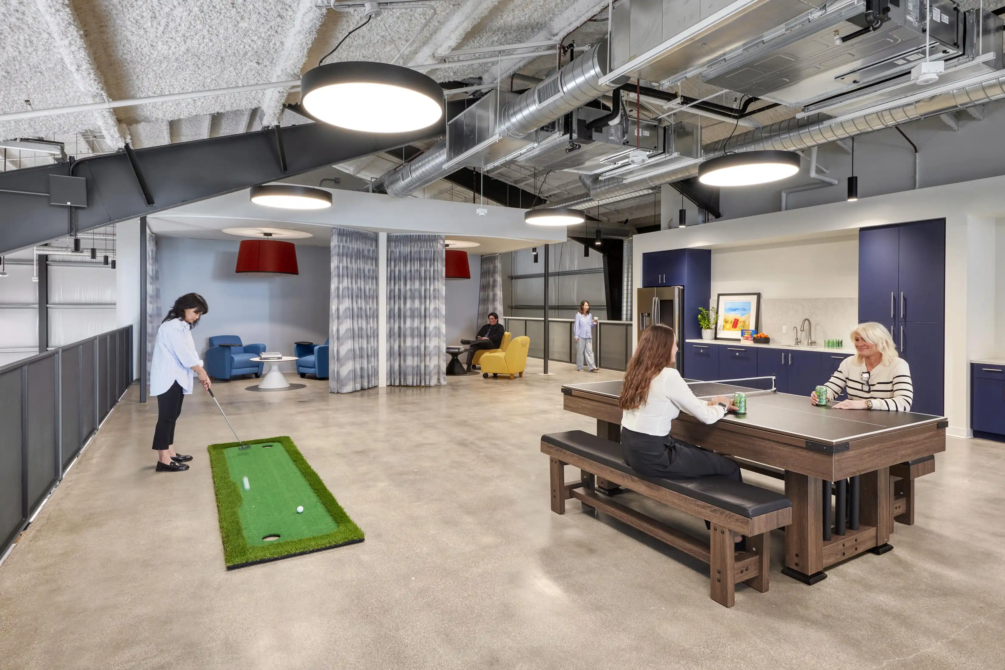 Open office space with a woman playing indoor golf on a mini green. Two women at a long table are conversing. Relaxed, modern atmosphere with cozy seating.