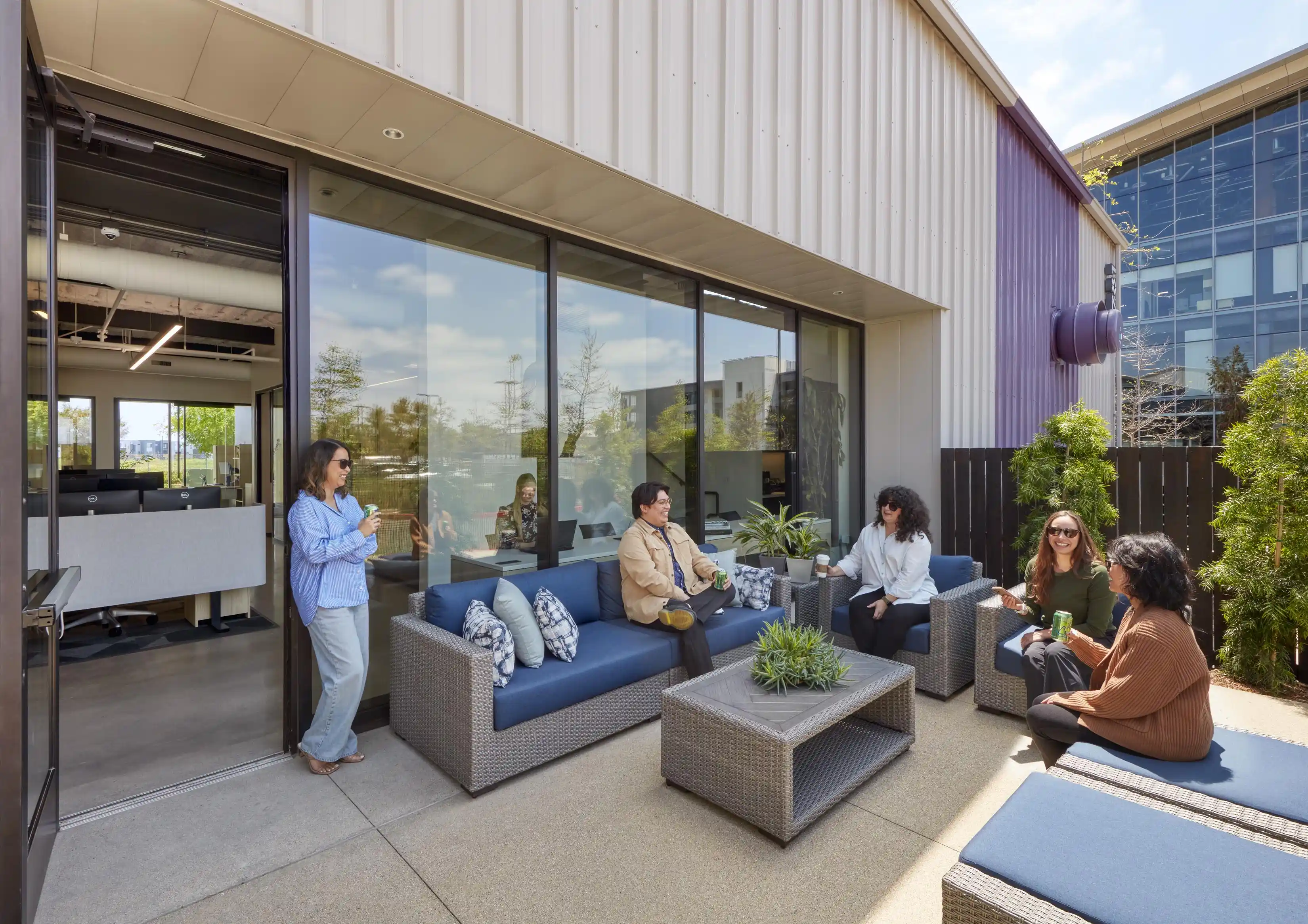 Five people are gathered on a modern patio with blue cushioned furniture, conversing and enjoying drinks. The setting is relaxed and sunny.