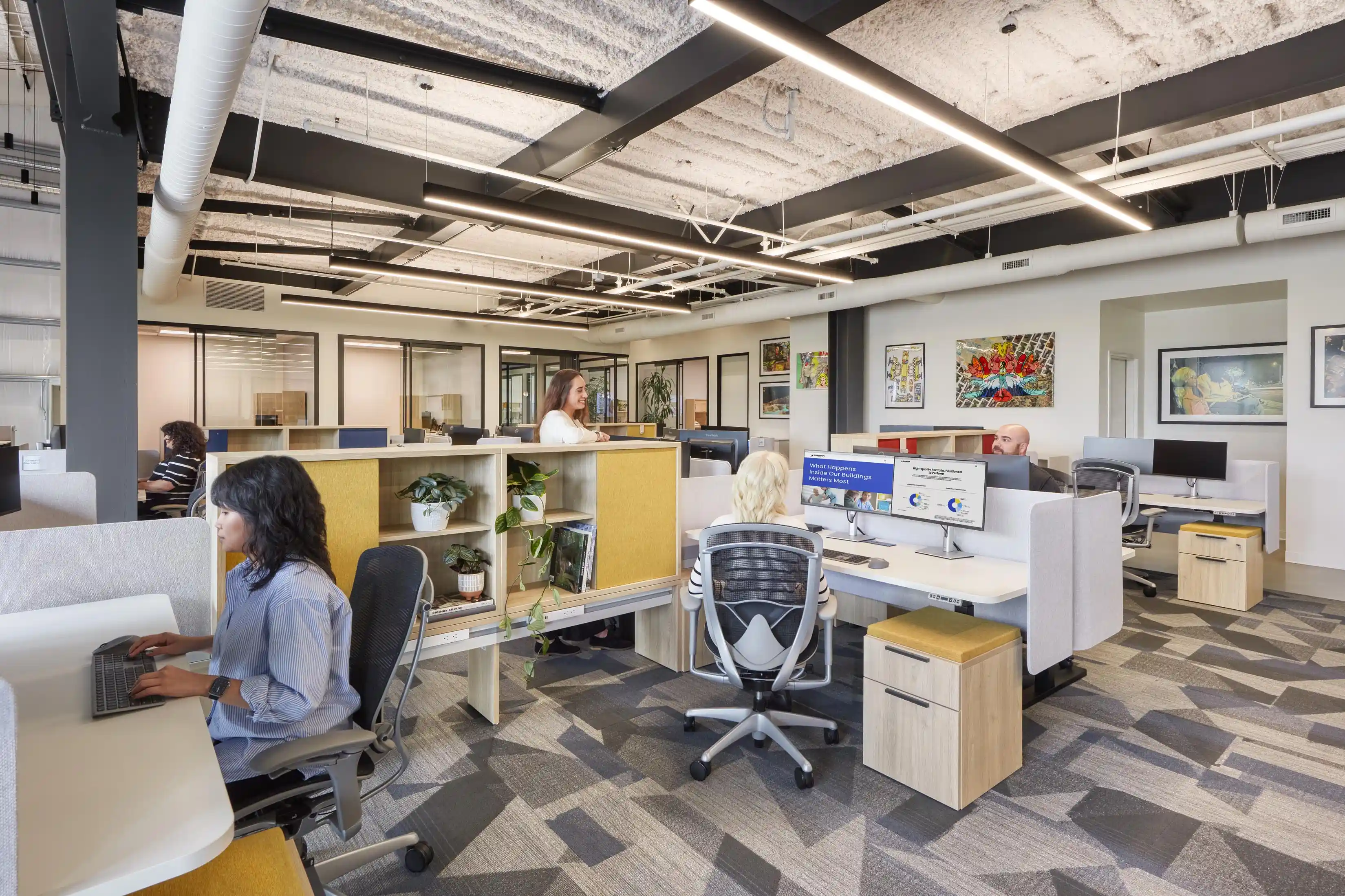 Open-plan office space with modern design, featuring people working at desks with computers. Bright lighting, patterned carpet, and colorful wall art create a vibrant atmosphere.