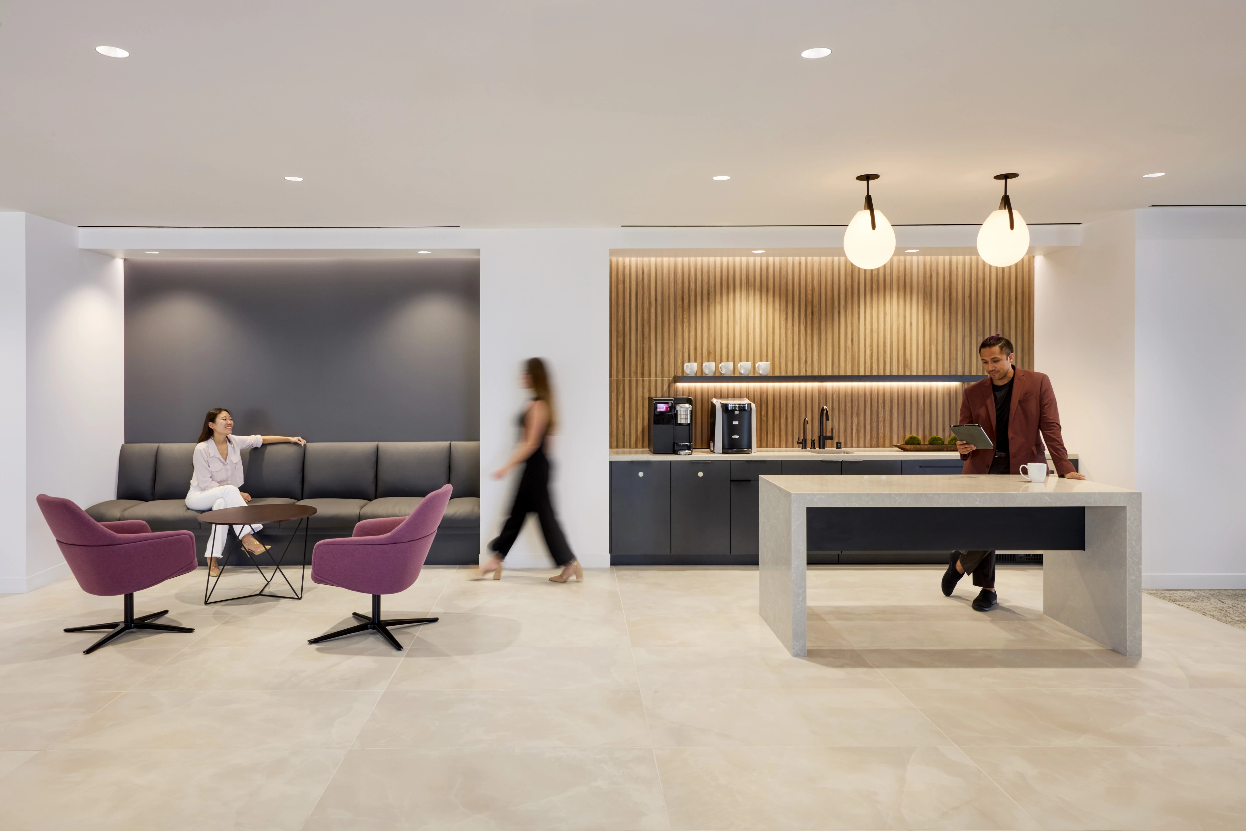 Modern office lounge with a relaxed atmosphere. A woman sits on a gray sofa, a man stands by a coffee bar with pendant lights, and a person walks by.