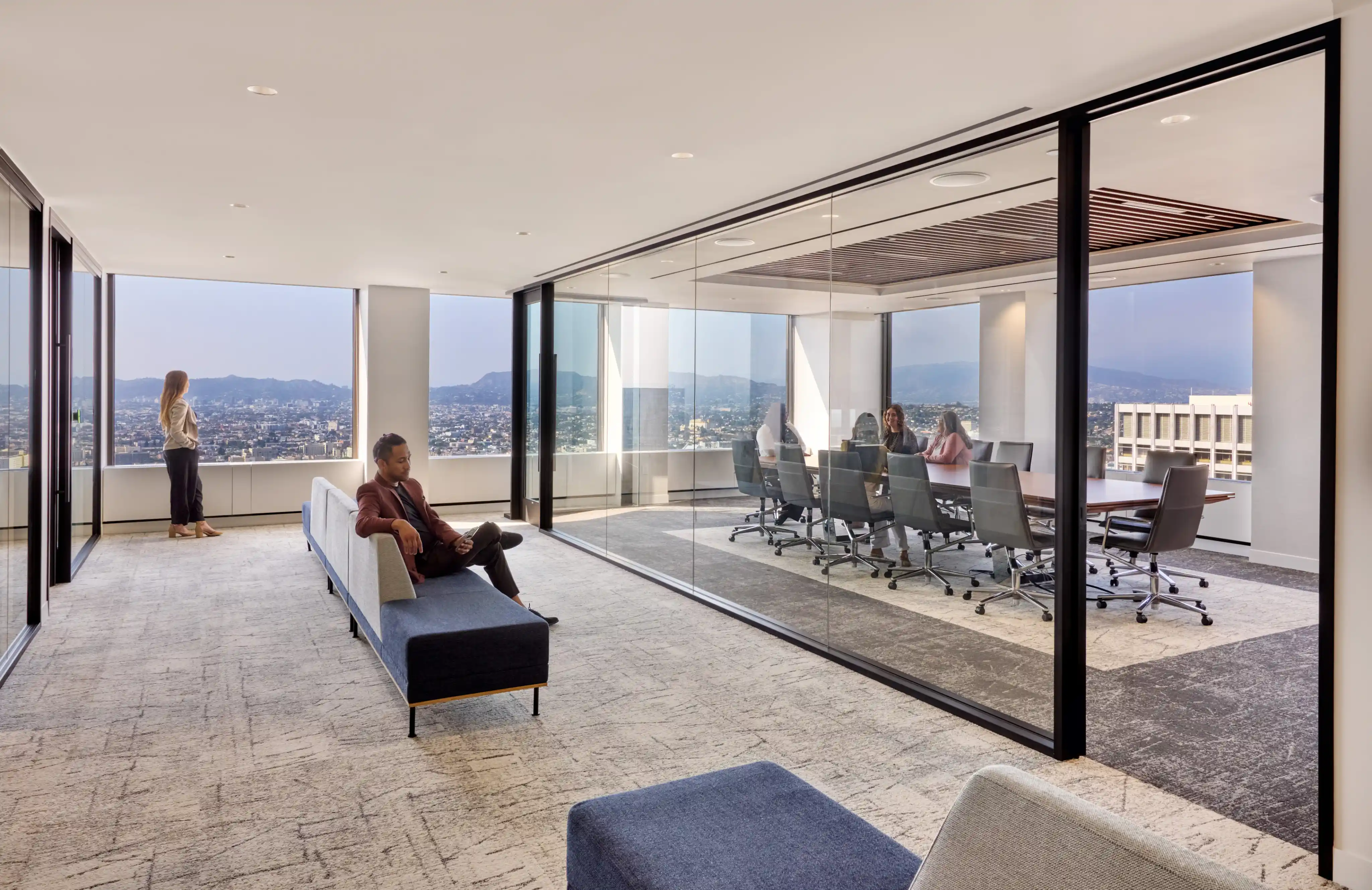 Modern office with floor-to-ceiling windows showing a city view. People sit in a glass-walled conference room and others are in the lounge area, creating a professional, serene atmosphere.