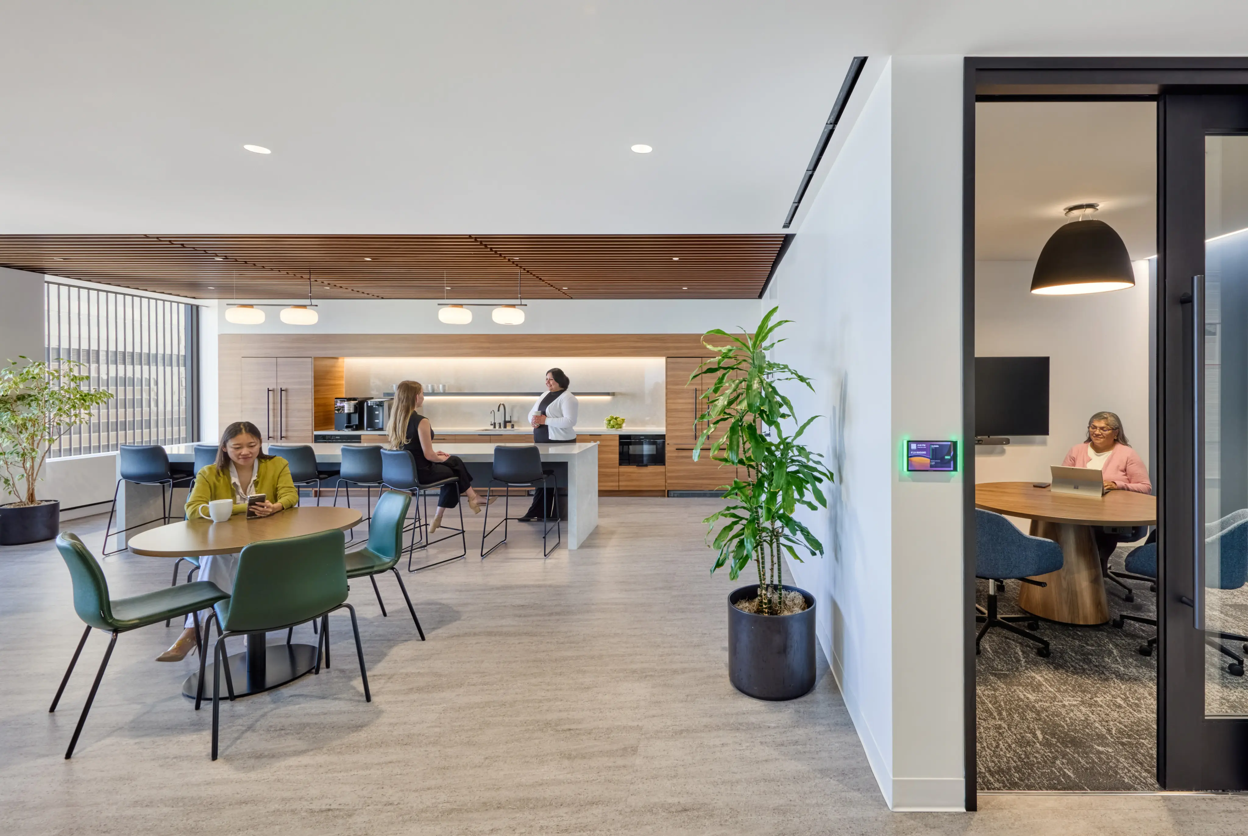 Modern office with a lounge; people work at tables and a coffee bar. A person in a conference room, glass walls, natural light, and plants enhance the serene ambiance.