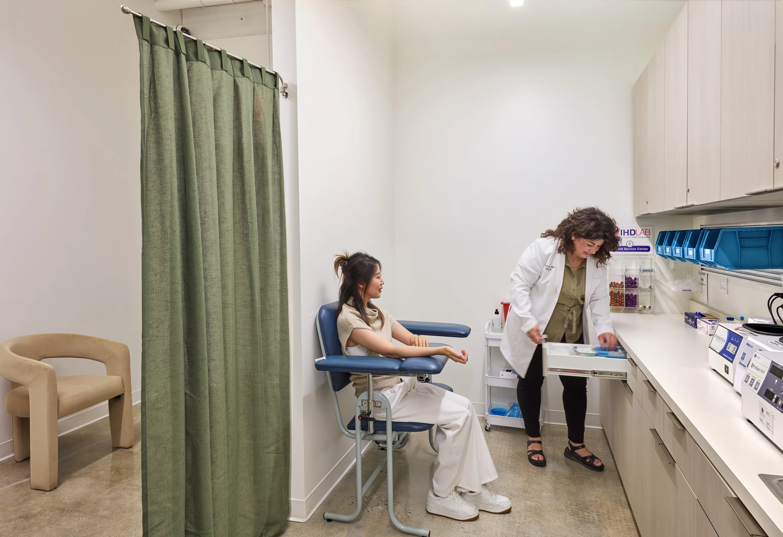 Doctor in a white coat prepares medical supplies while a seated woman waits in a clinic room. The setting is clean and clinical, conveying a sense of care.