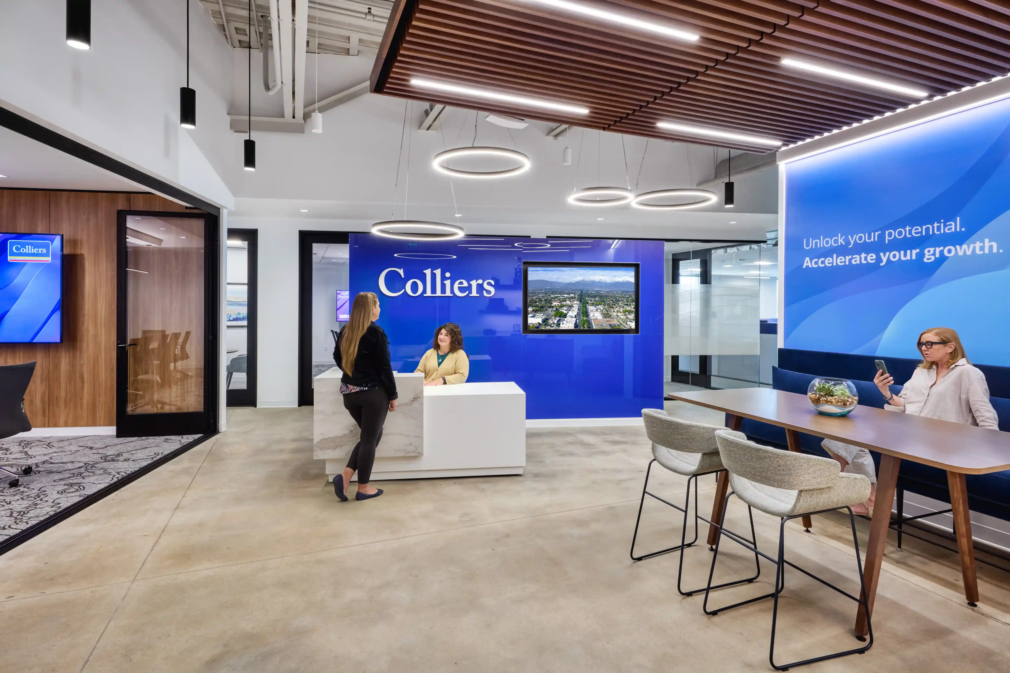 Modern office reception with two women interacting at a white desk. A large screen and "Colliers" logo are on the blue wall. Another woman is seated at a wooden table with chairs, viewing a phone. The atmosphere is professional and welcoming, with circular