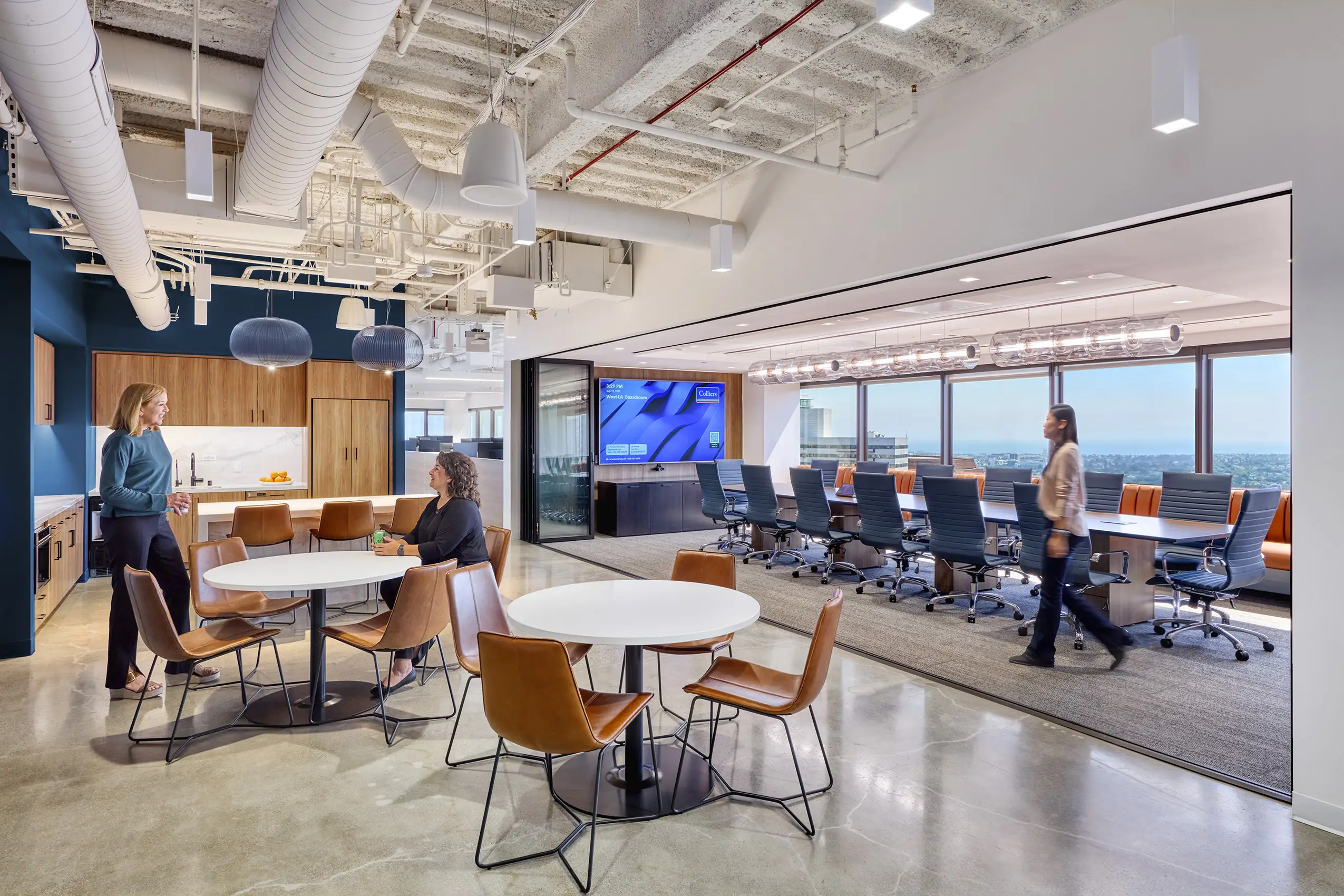 Modern office space with an open seating area showing round tables and brown chairs. Two people converse near a kitchenette. A woman walks past a glass-walled conference room with a large screen and city views, creating a collaborative and professional atm