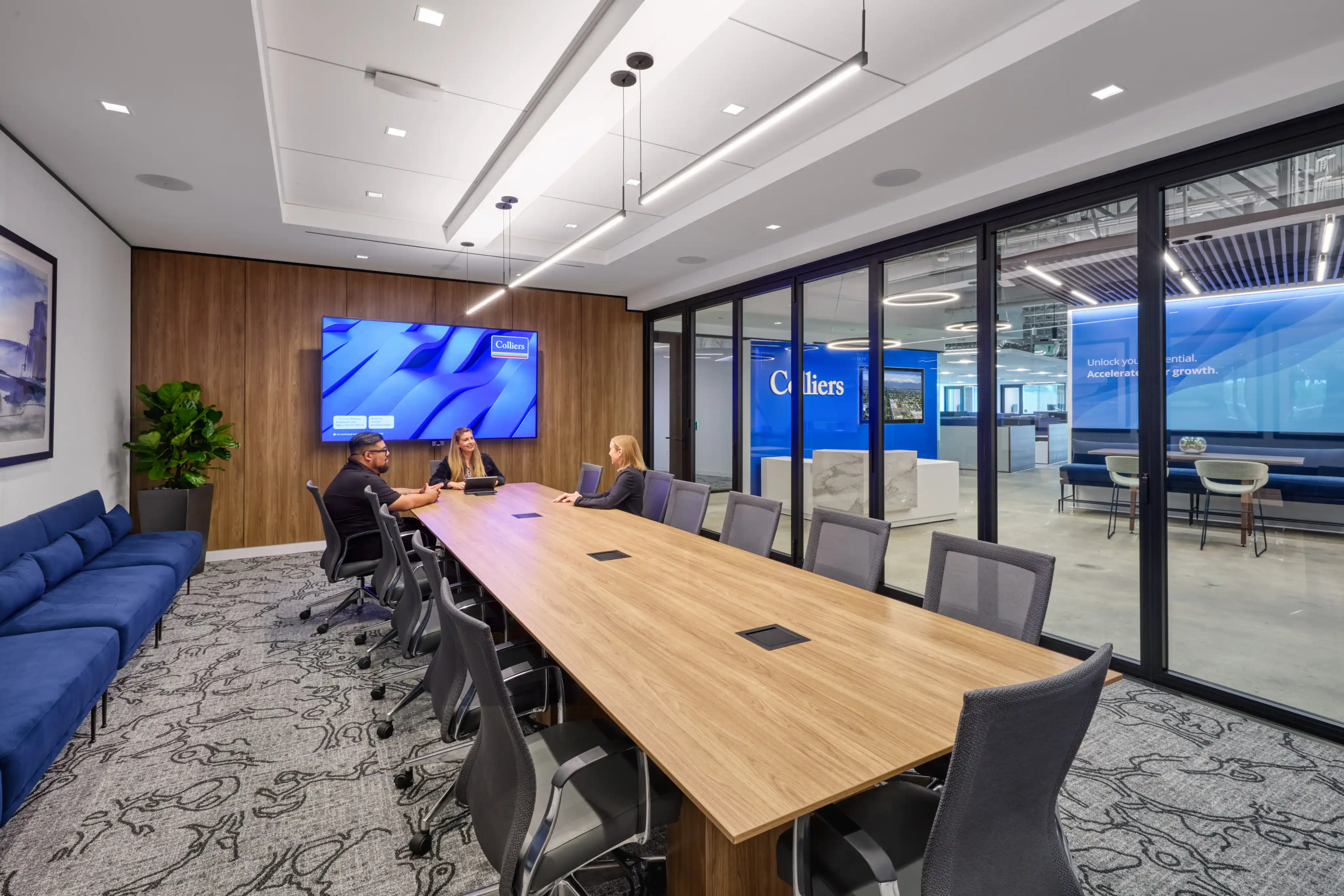 A modern conference room with three people sitting at a long wooden table, facing a large screen displaying a blue image. Glass walls reveal an adjacent office area.