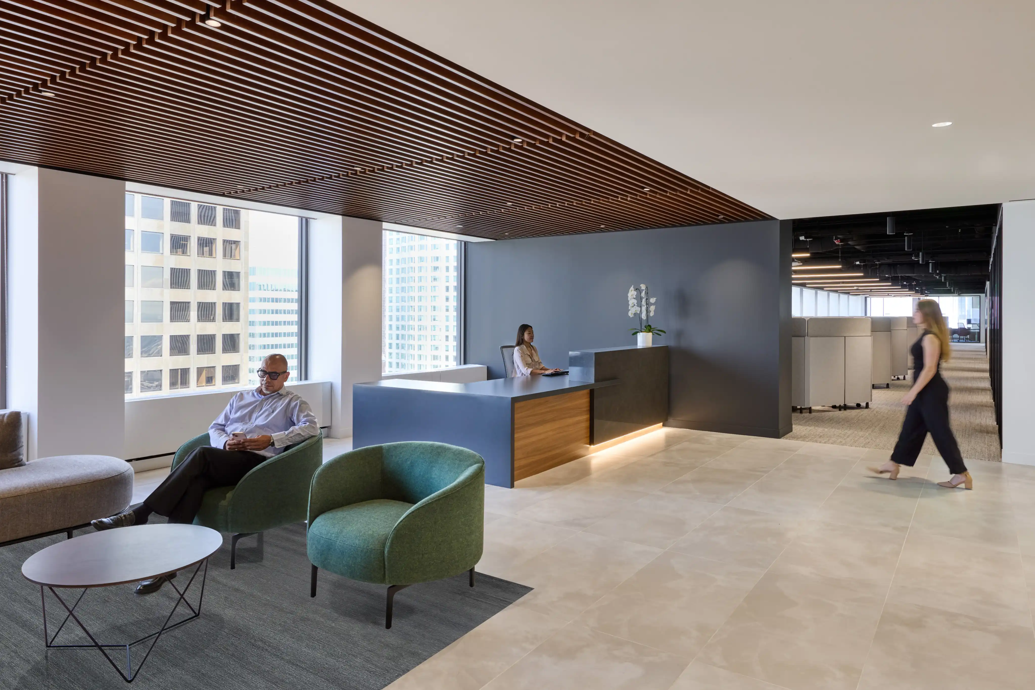 Modern office lobby with wooden slatted ceiling, blue reception desk, and seating area with green chairs. A person walks past, conveying a calm, professional atmosphere.