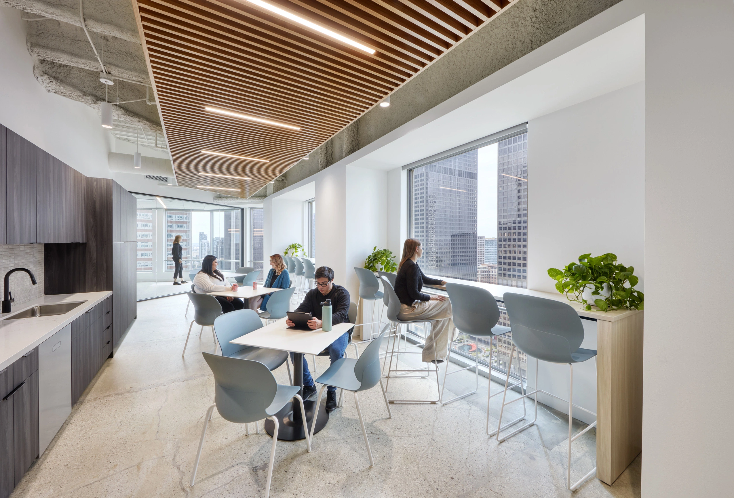 Modern office break room with wooden ceiling panels and large windows. People work and relax at tables and a counter, surrounded by plants, exuding a calm atmosphere.