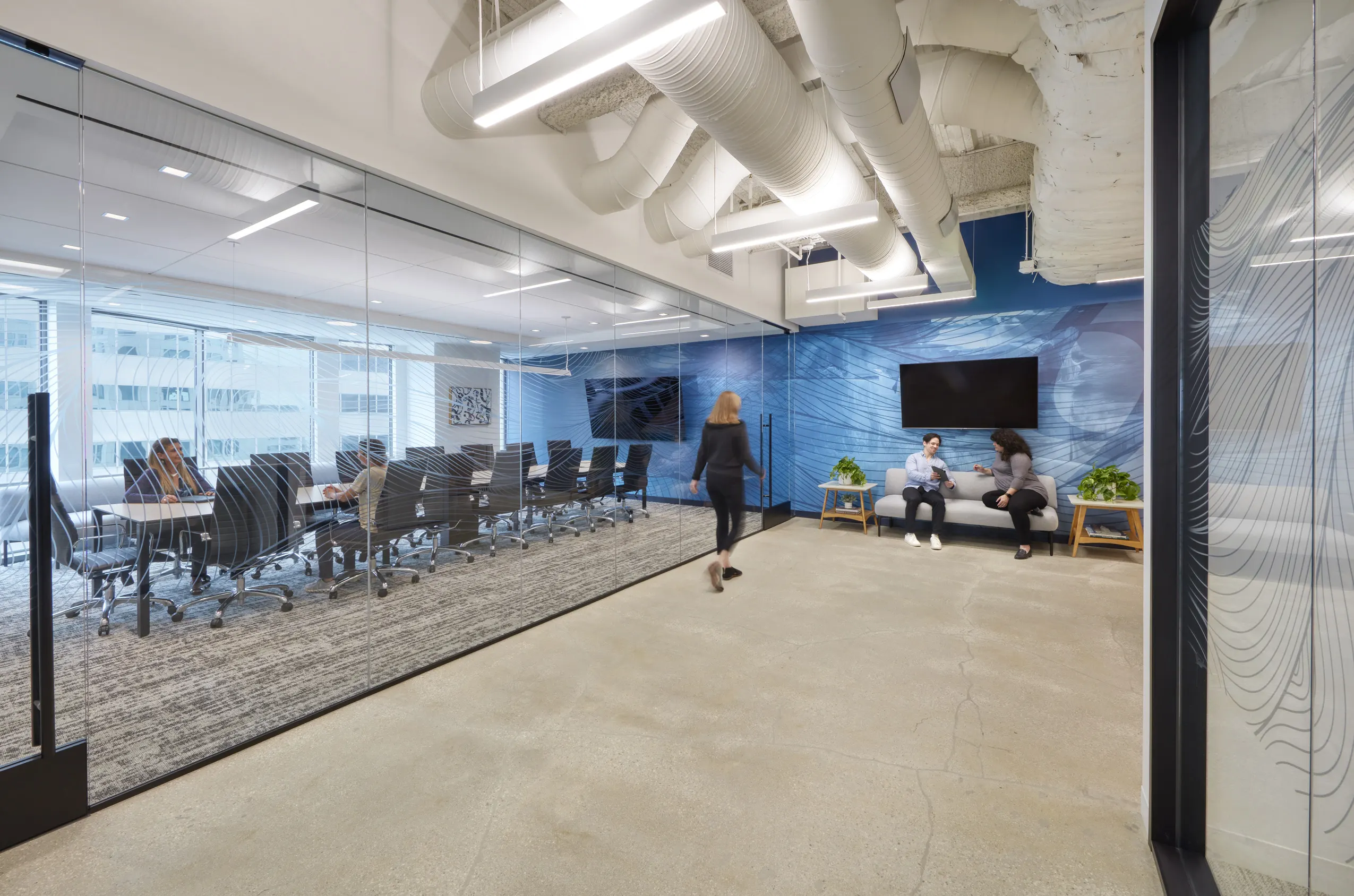 Modern office space featuring a glass-walled conference room with chairs, three people sitting on couches in the hallway, and a person walking by. Bright, clean, and professional.