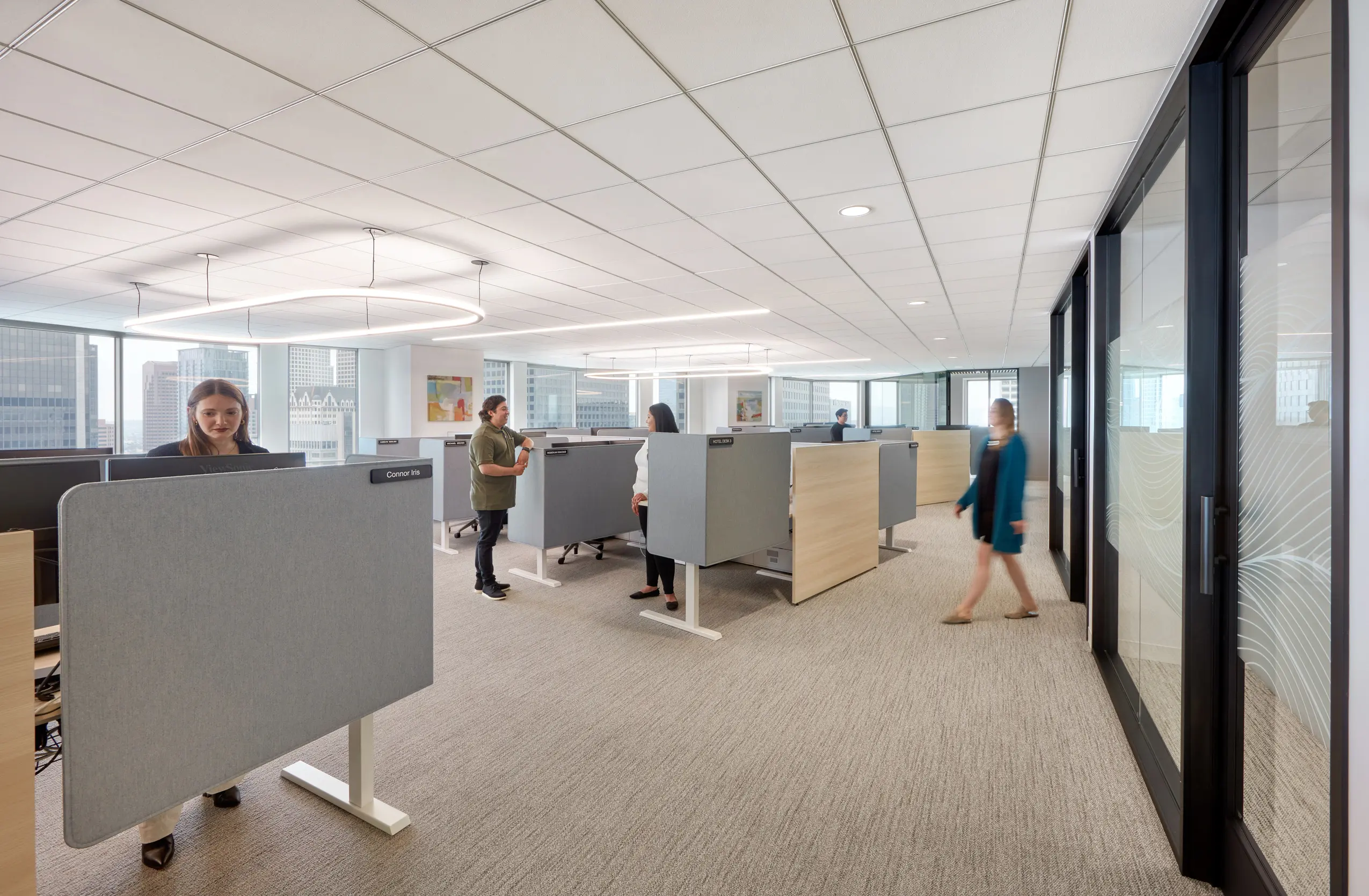 Modern office space with people working at standing desks separated by partitions. Glass walls and large windows offer a bright, collaborative atmosphere.