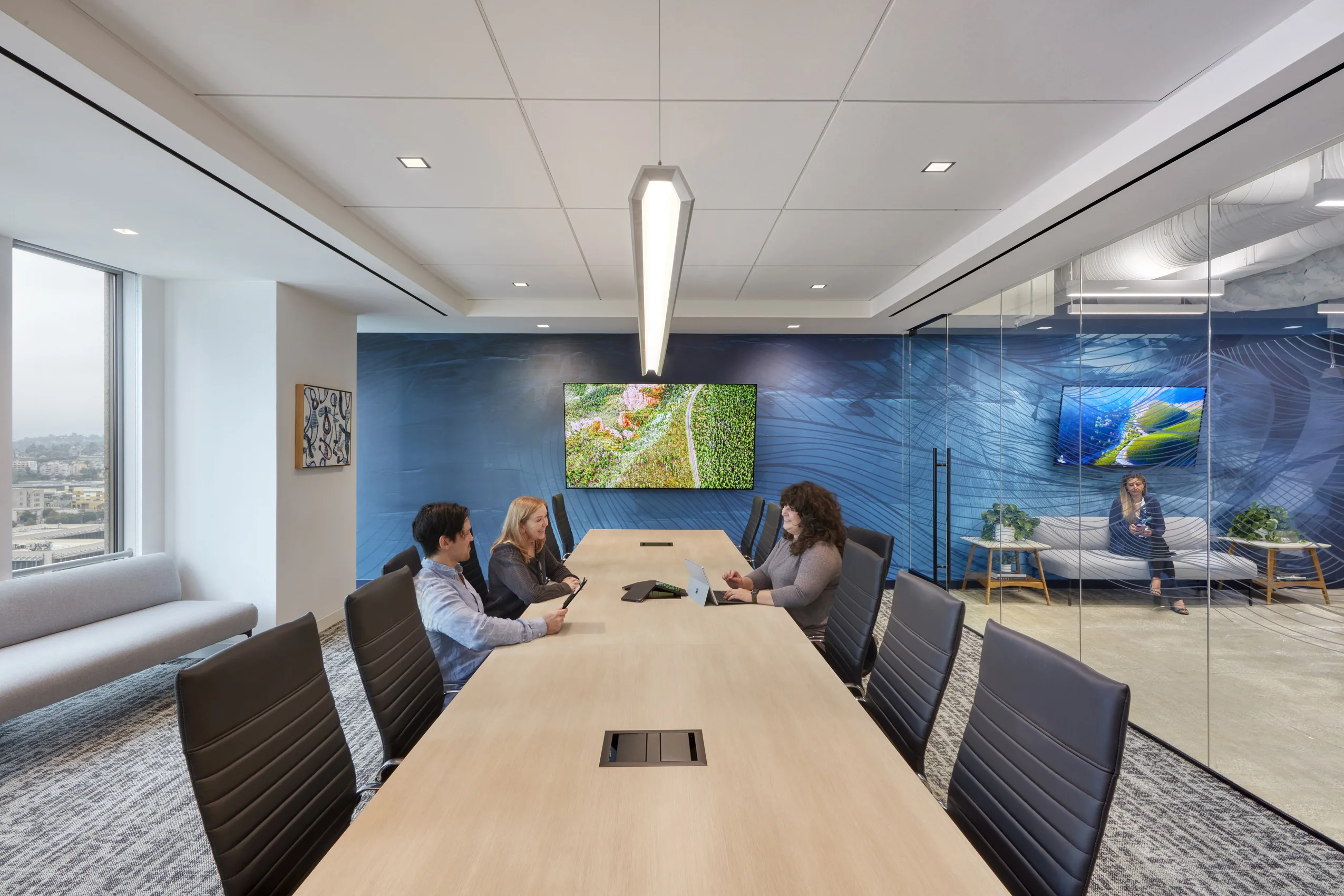 A modern, spacious conference room with a long wooden table and black chairs. Three people engage in discussion. The blue accent wall features screens displaying nature scenes. A glass wall divides the area, where another person sits on a sofa, creating a 