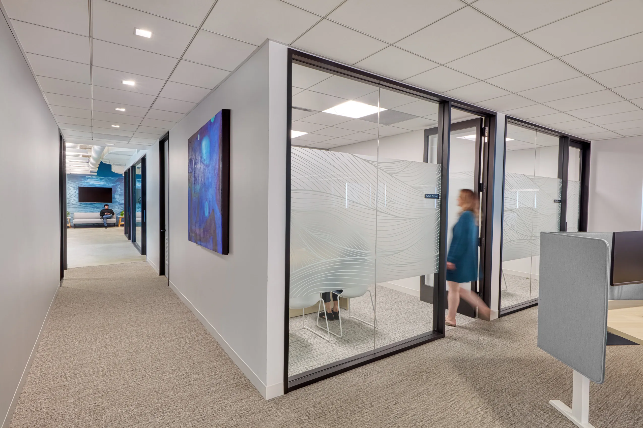 Modern office hallway with glass-walled meeting rooms on the right, decorated with abstract frosted patterns. A person walks past in a blue outfit.