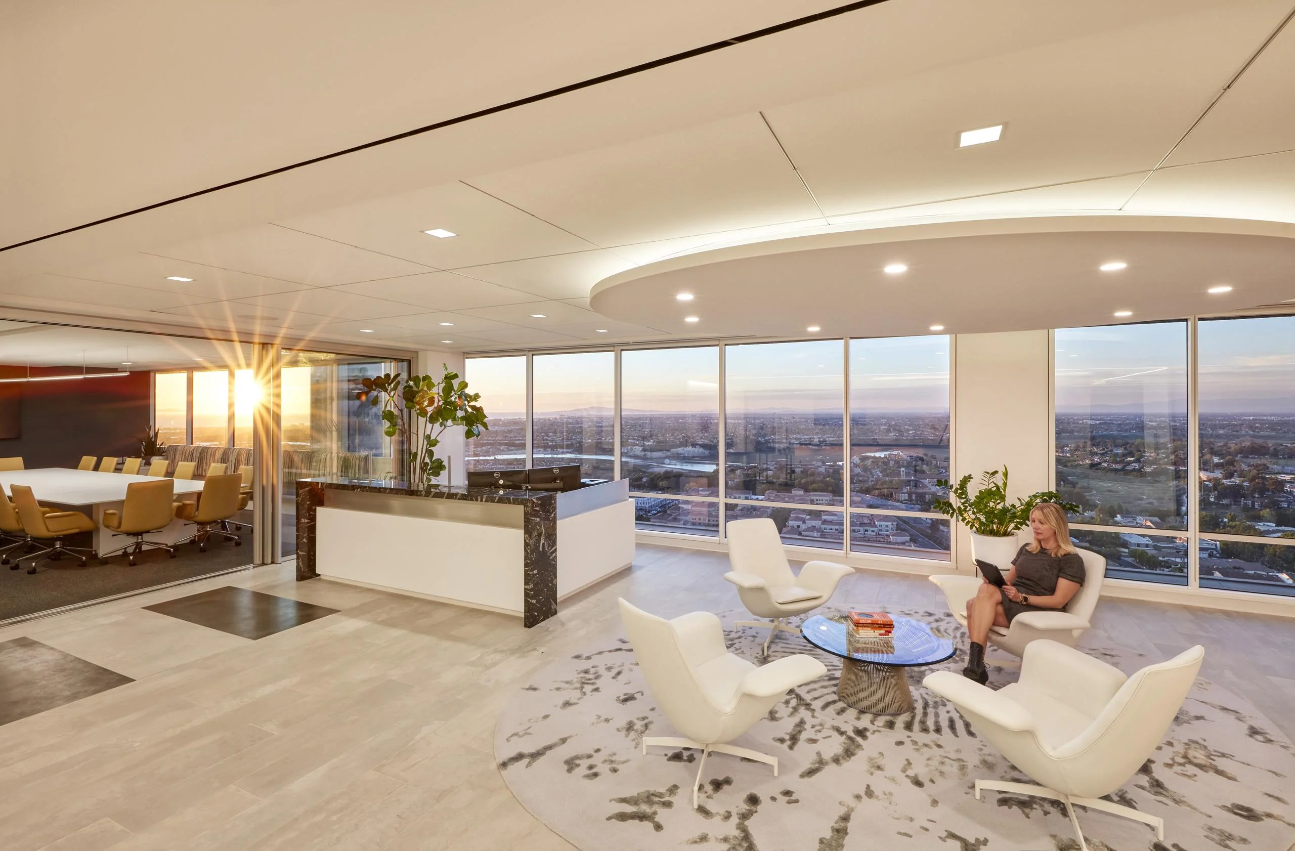 Luxurious office lounge with white chairs around a glass table, large windows showing a sunset city view, and a person seated, reading.