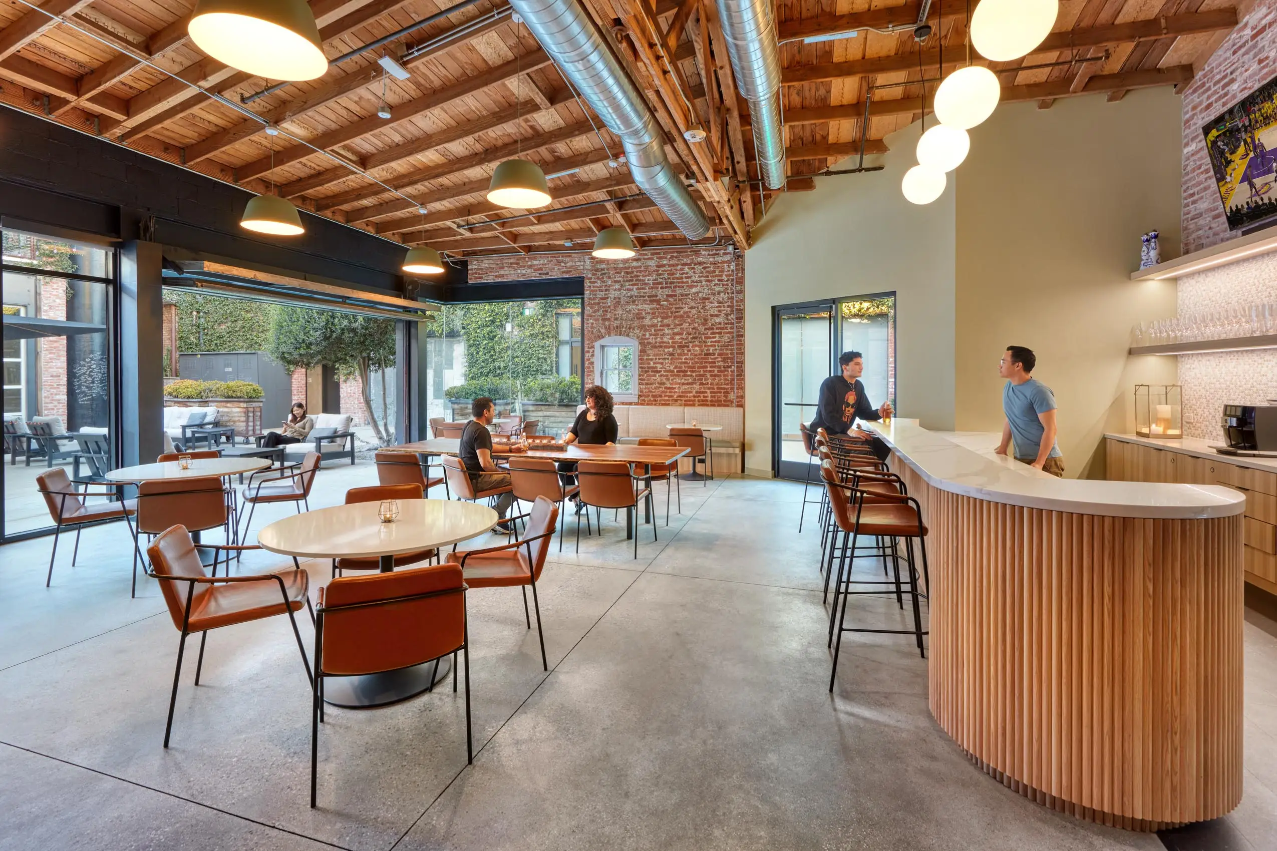 Modern cafe with brick walls, wooden ceiling, and industrial lighting. People sit and chat at round tables and a curved bar, creating a relaxed atmosphere.