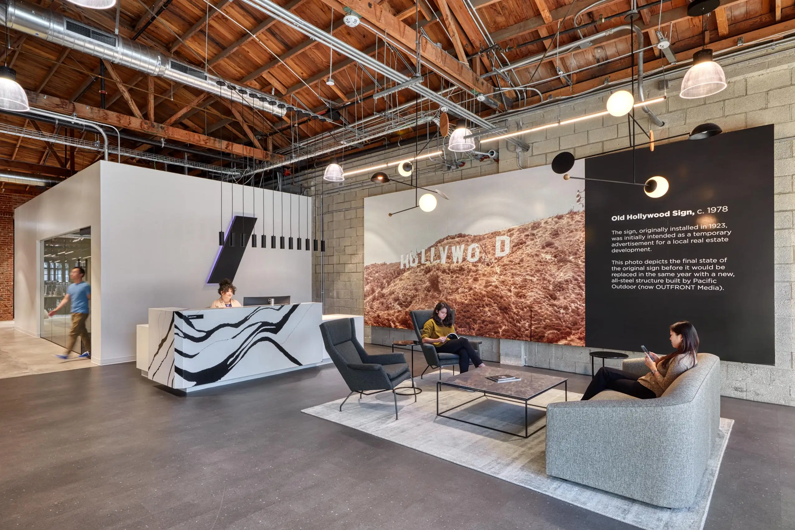 Modern office lobby with exposed wooden ceiling and industrial lights. Features a reception desk, seating area, and a mural of the Hollywood sign wall art.