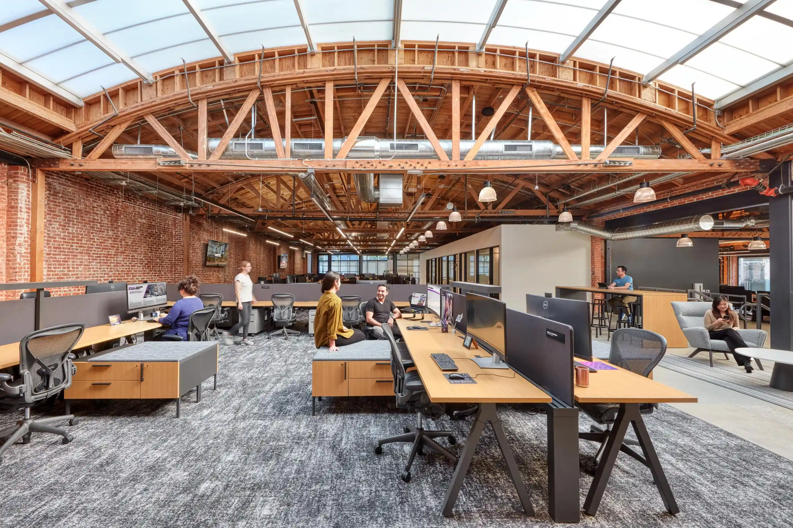 Modern open-office space with exposed brick walls and wooden beams. People work at desks and lounge in seating areas. Bright, airy atmosphere.
