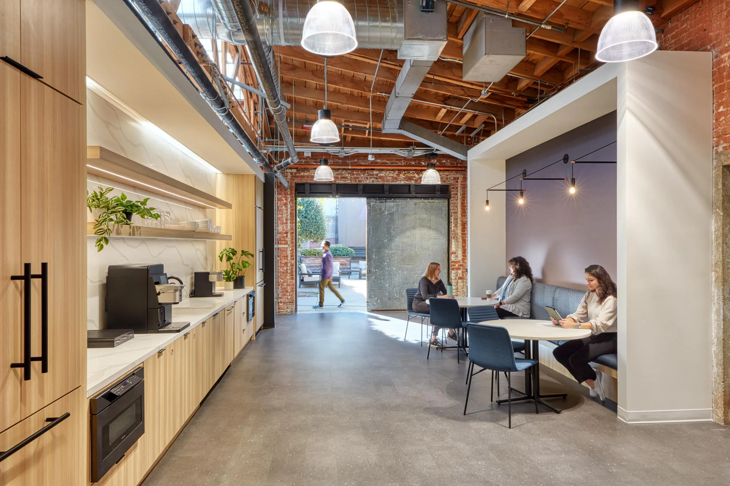 Modern office kitchen with wooden cabinets and plants, adjacent to a seating area with tables. A person walks by an open industrial-style door.