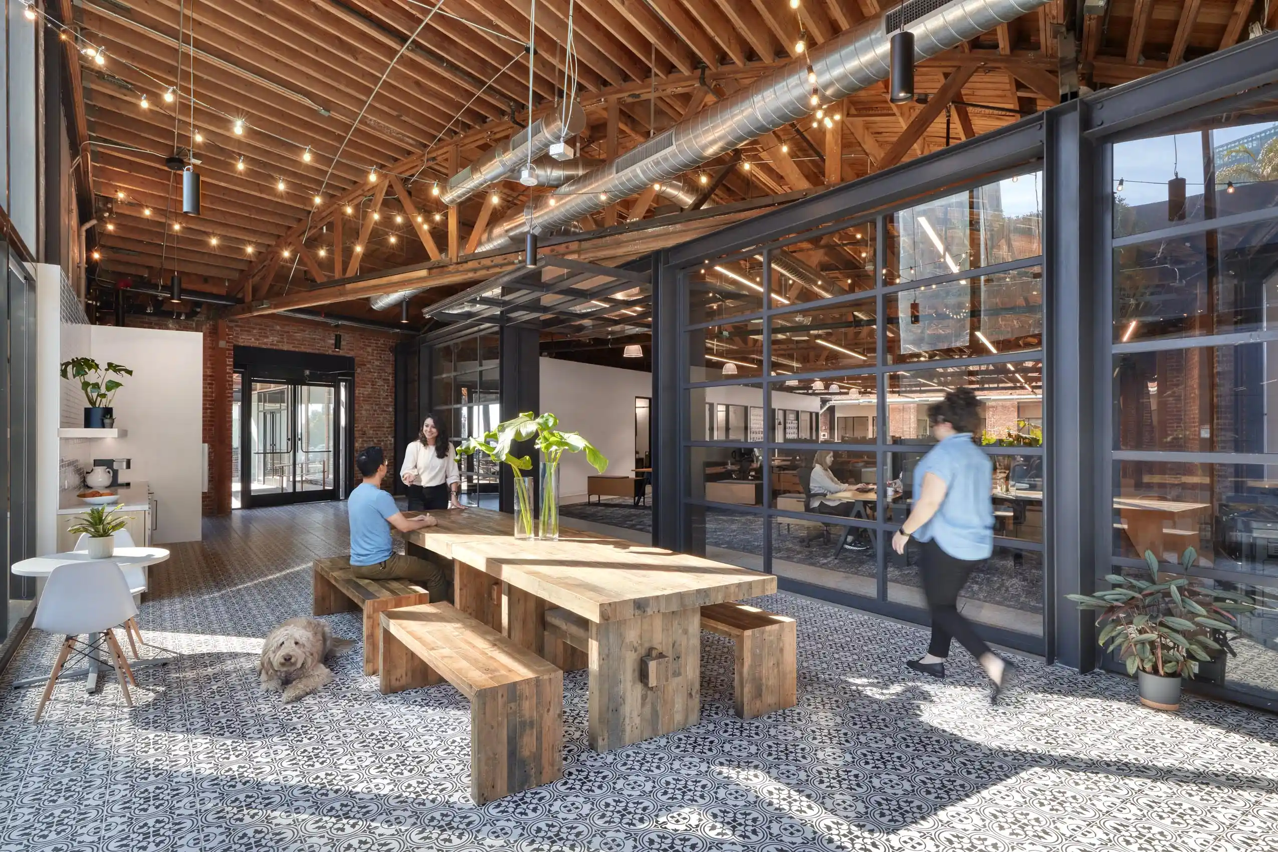 Spacious office interior with a wooden ceiling and glass walls. A large wooden table is centered with people and a dog. The atmosphere feels open and collaborative.