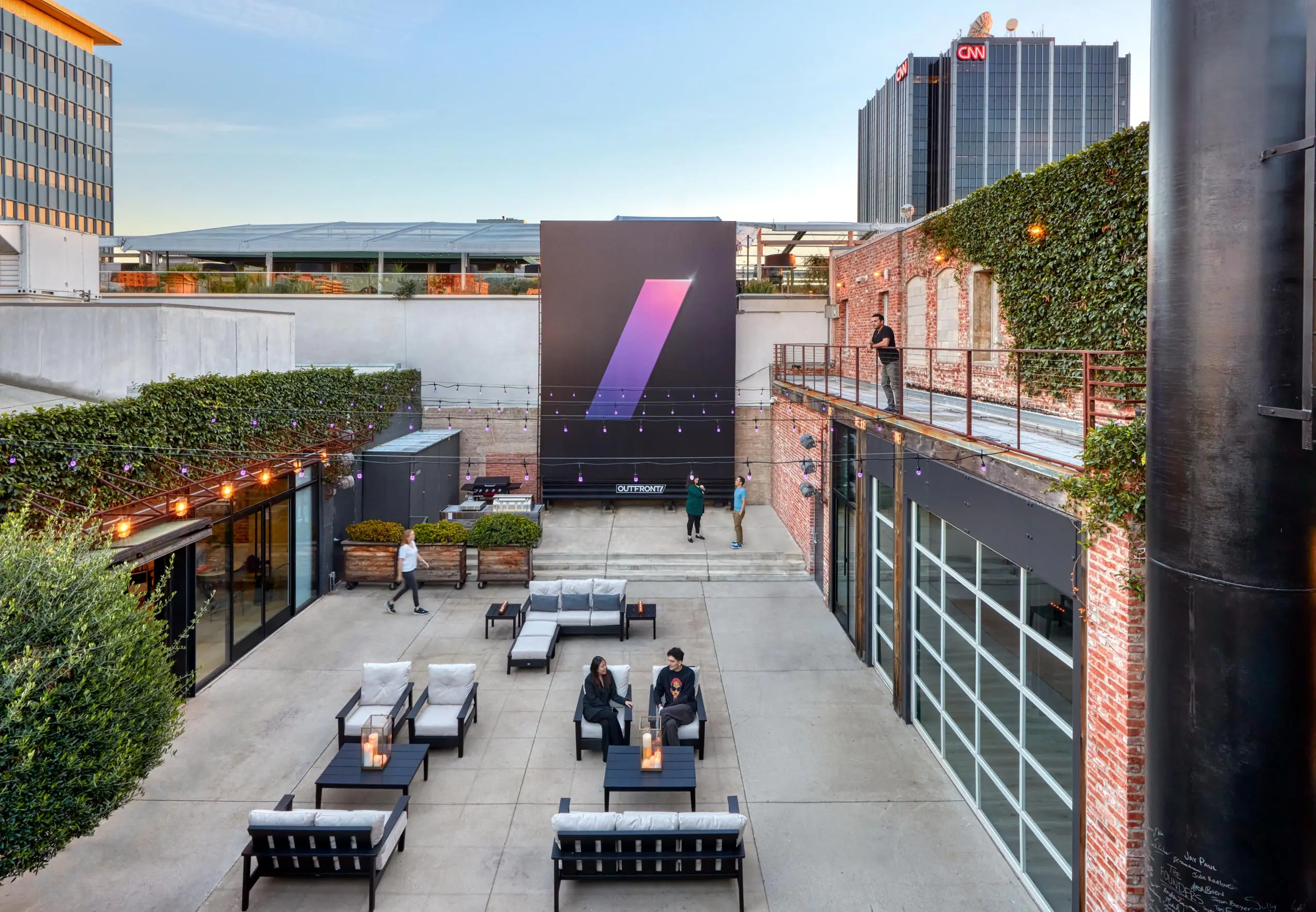Modern outdoor patio with sofas and tables on a concrete floor. Brick walls adorned with greenery. Large mural. Relaxed, urban atmosphere.