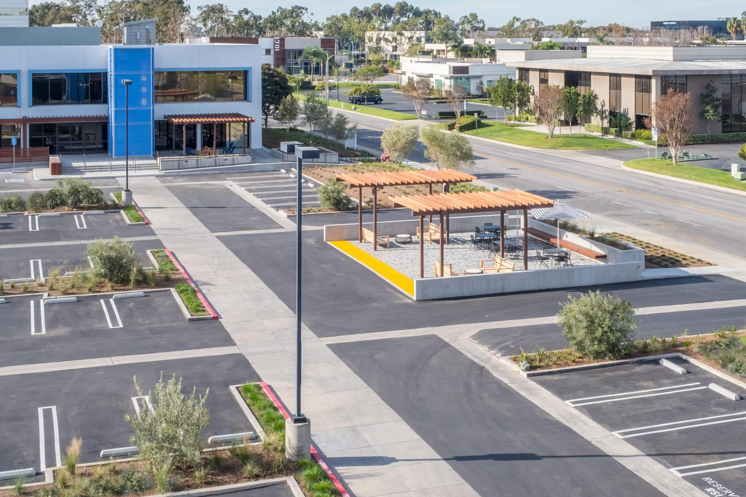 Aerial view of a nearly empty parking lot with a modern building on the left. There are two pergolas with outdoor seating in the center, flanked by trees and a road. The scene is calm and suburban.