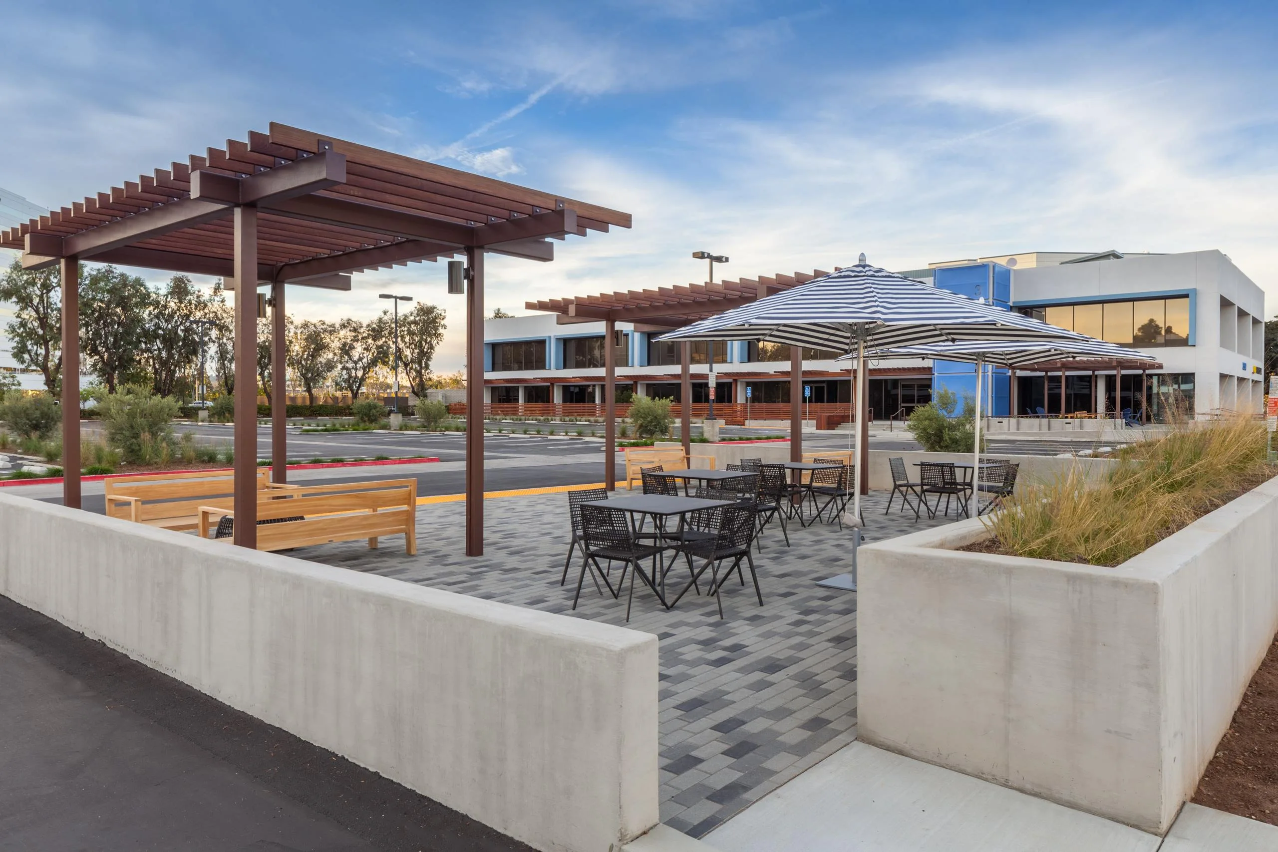 Outdoor seating area with wooden pergolas and black metal tables under striped umbrellas. Modern buildings and landscaped greenery in the background.