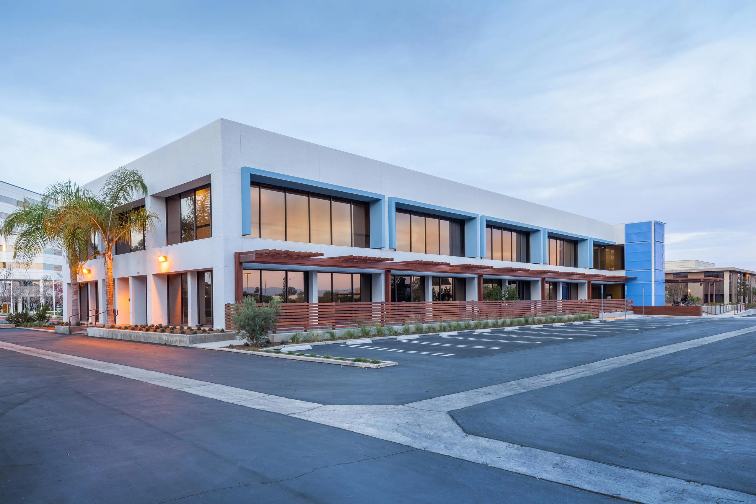 Modern two-story office building with large tinted windows, surrounded by palm trees and an empty parking lot, under a clear, tranquil sky.