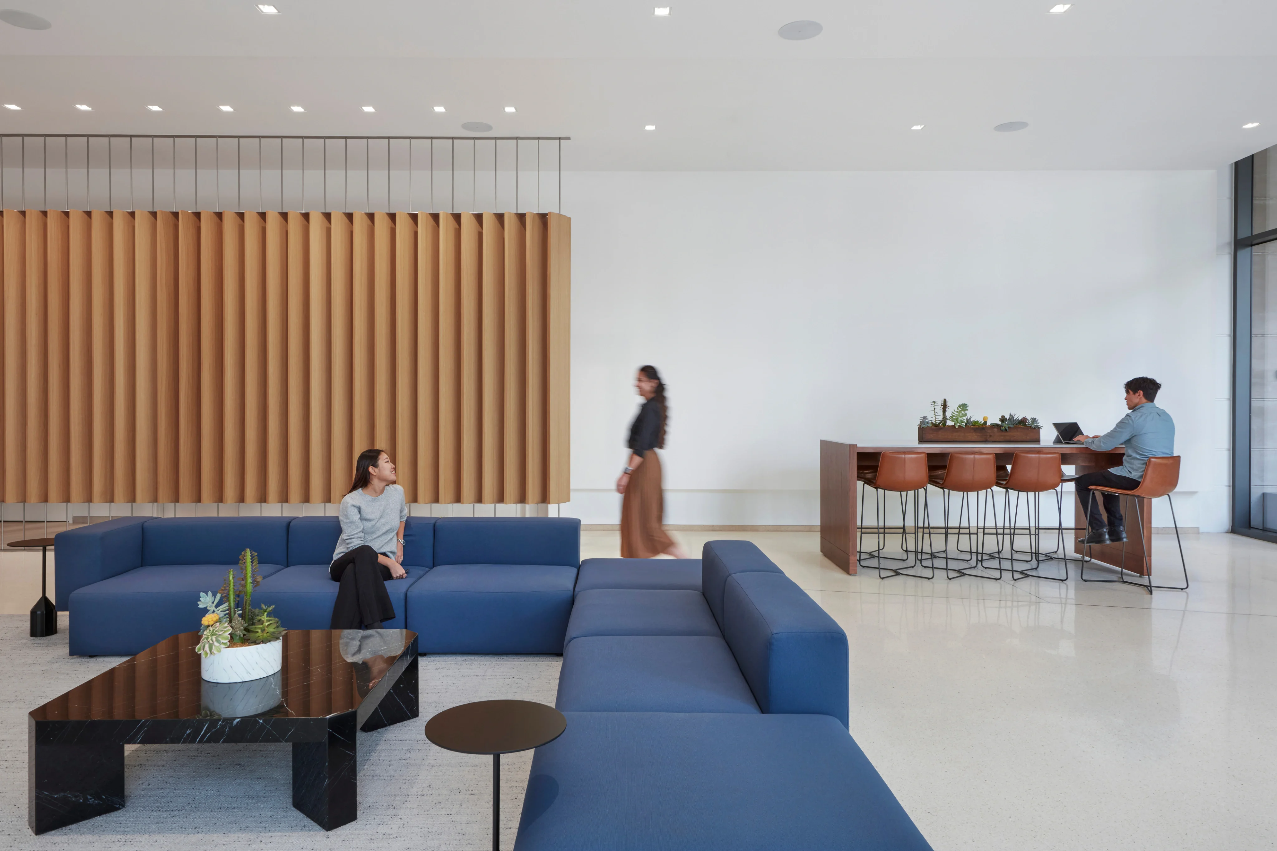 Modern office lounge with blue sofas and a black table. A woman sits on the sofa, another walks by, and a man works on a laptop at a bar table. Emphasizes a calm, professional atmosphere.
