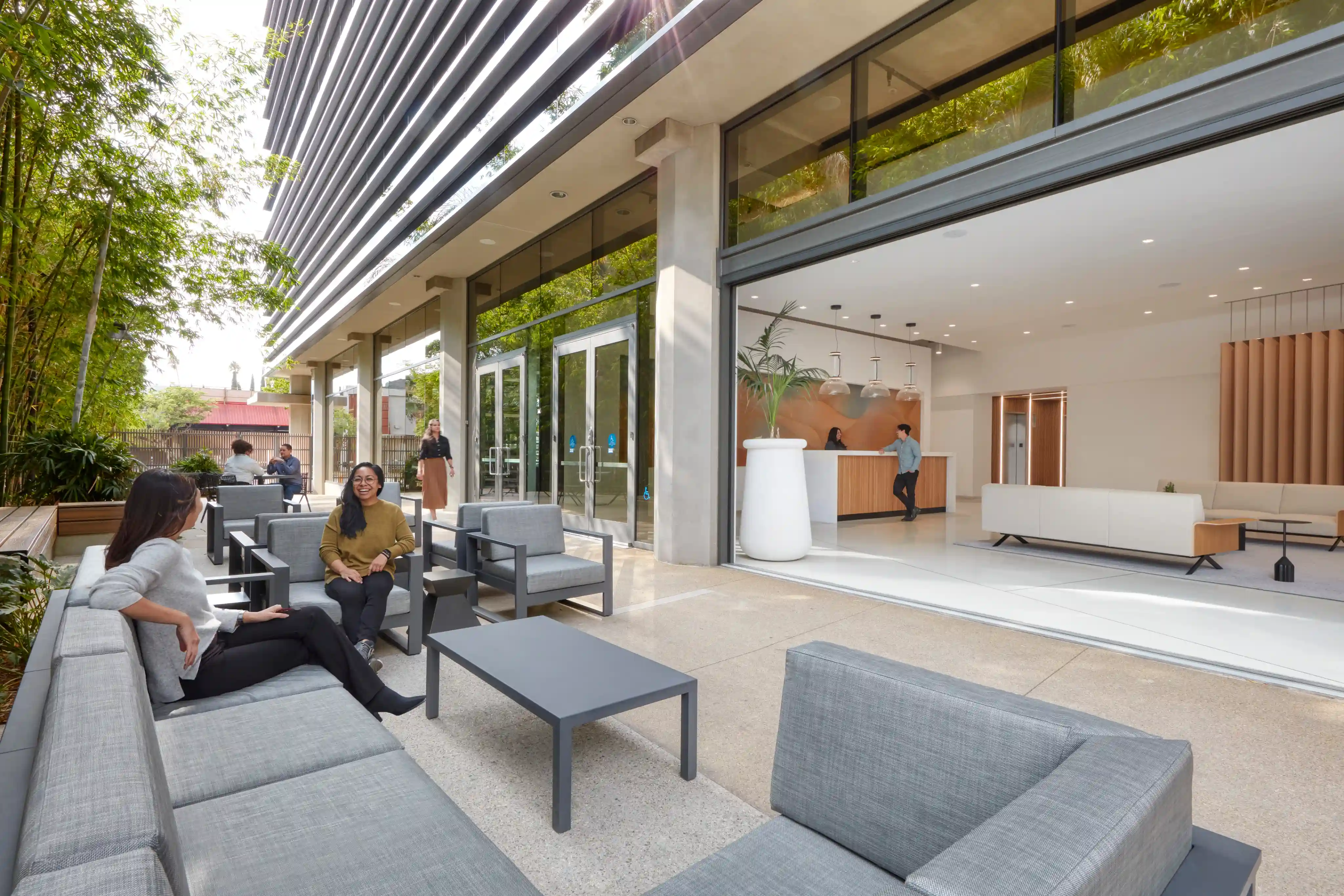 Modern office entrance with glass doors and a patio. People sit on gray outdoor furniture, talking. Inside, a person at a desk greets visitors. Bright, open atmosphere.