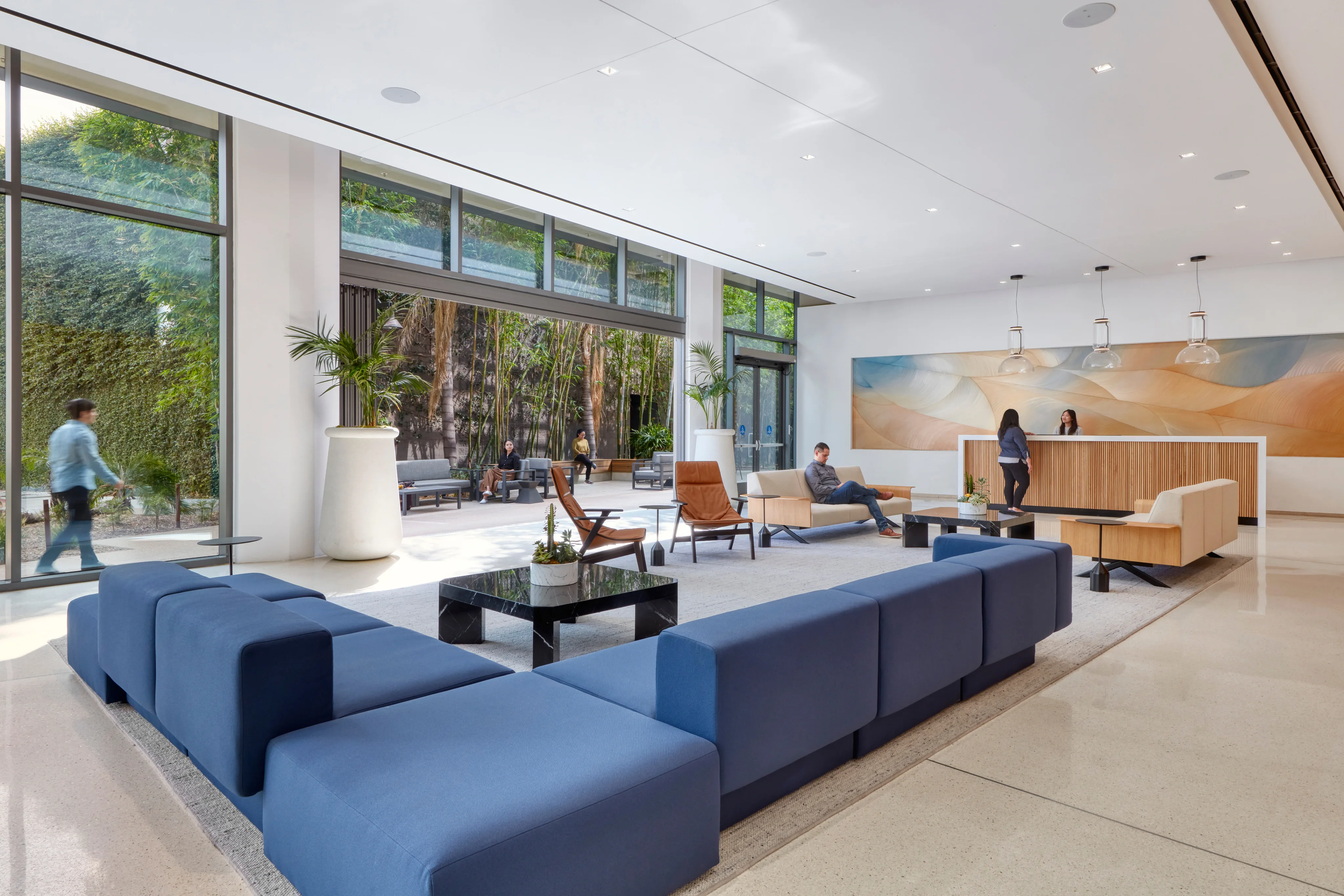 Modern lobby with blue sectional sofas, large windows showcasing greenery, and people conversing. A reception desk and abstract art add elegance.