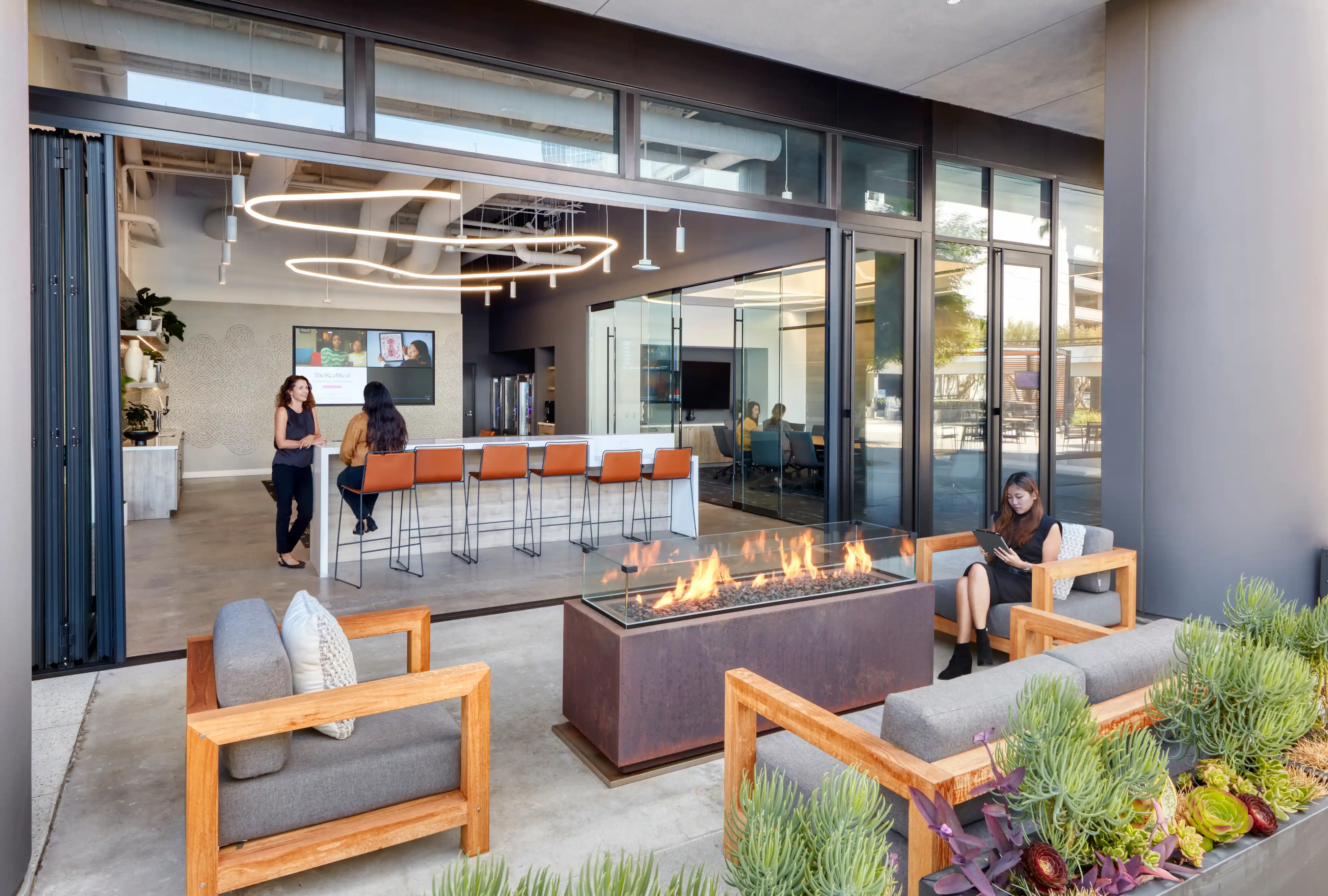 Modern indoor-outdoor lounge with a sleek fire pit and wooden chairs. Open glass doors reveal a meeting space with people and stylish lighting. Relaxed vibe.