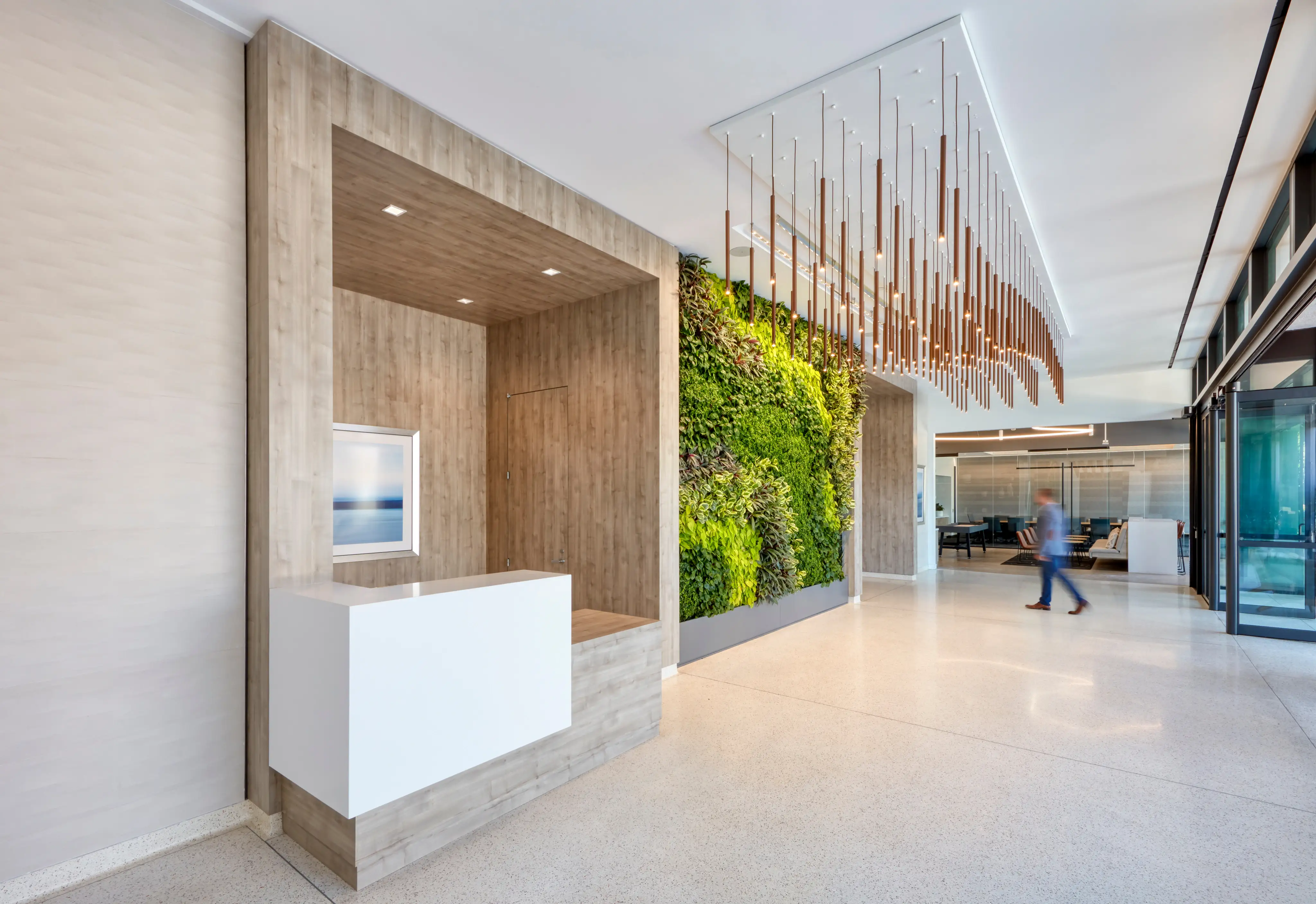 Modern lobby with a wooden reception desk, vertical green plant wall, and hanging linear light fixture. A person walks past, adding motion.