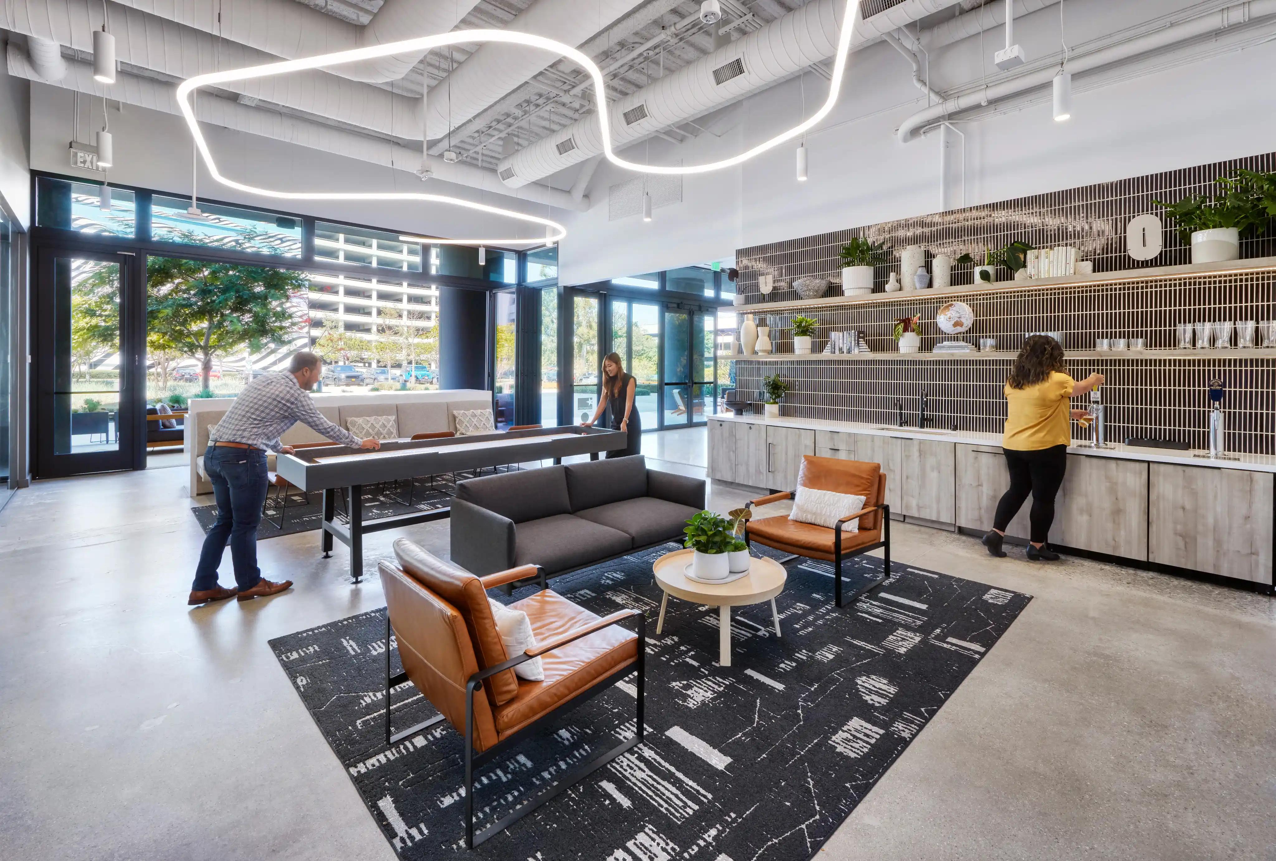 Modern lounge with open ceiling and wavy lights. People play table tennis and use the kitchenette. Brown chairs, black rug, and potted plants add warmth.