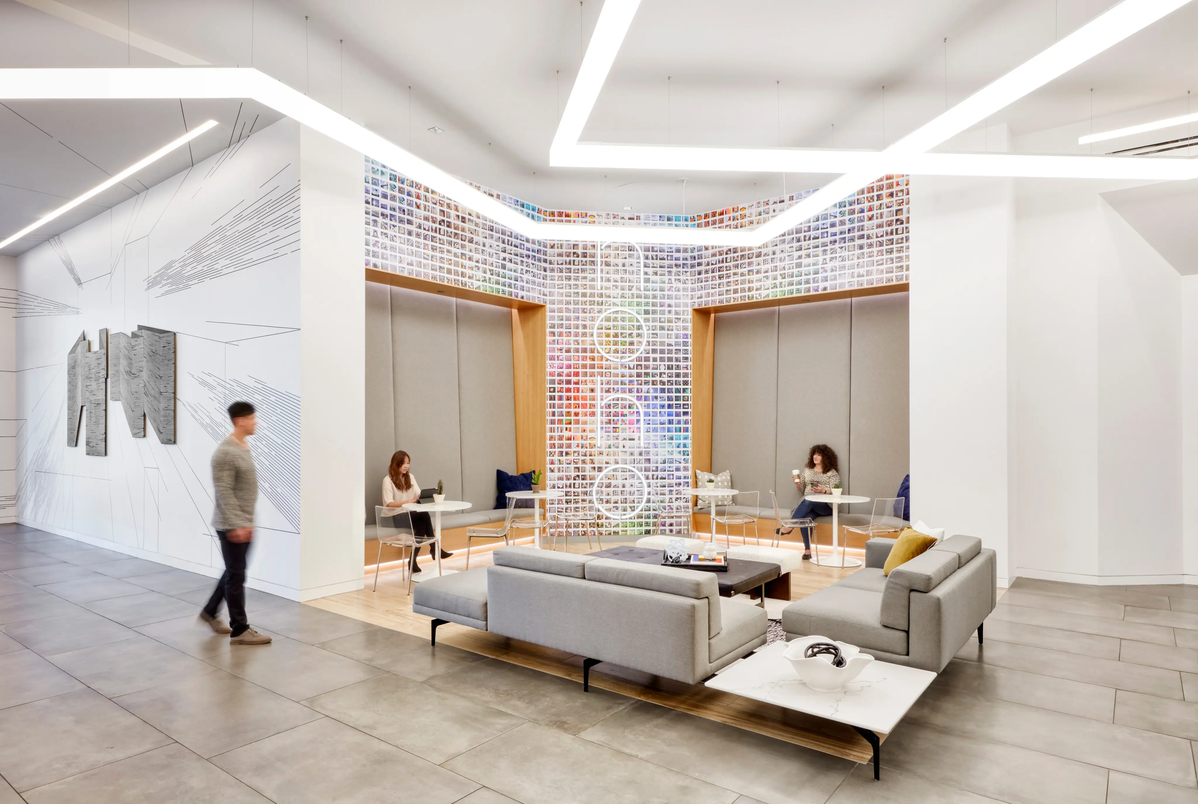 Modern office lounge with gray sofas and vibrant wall art. Three people work at small tables. Bright overhead lighting adds a sleek, open feel.