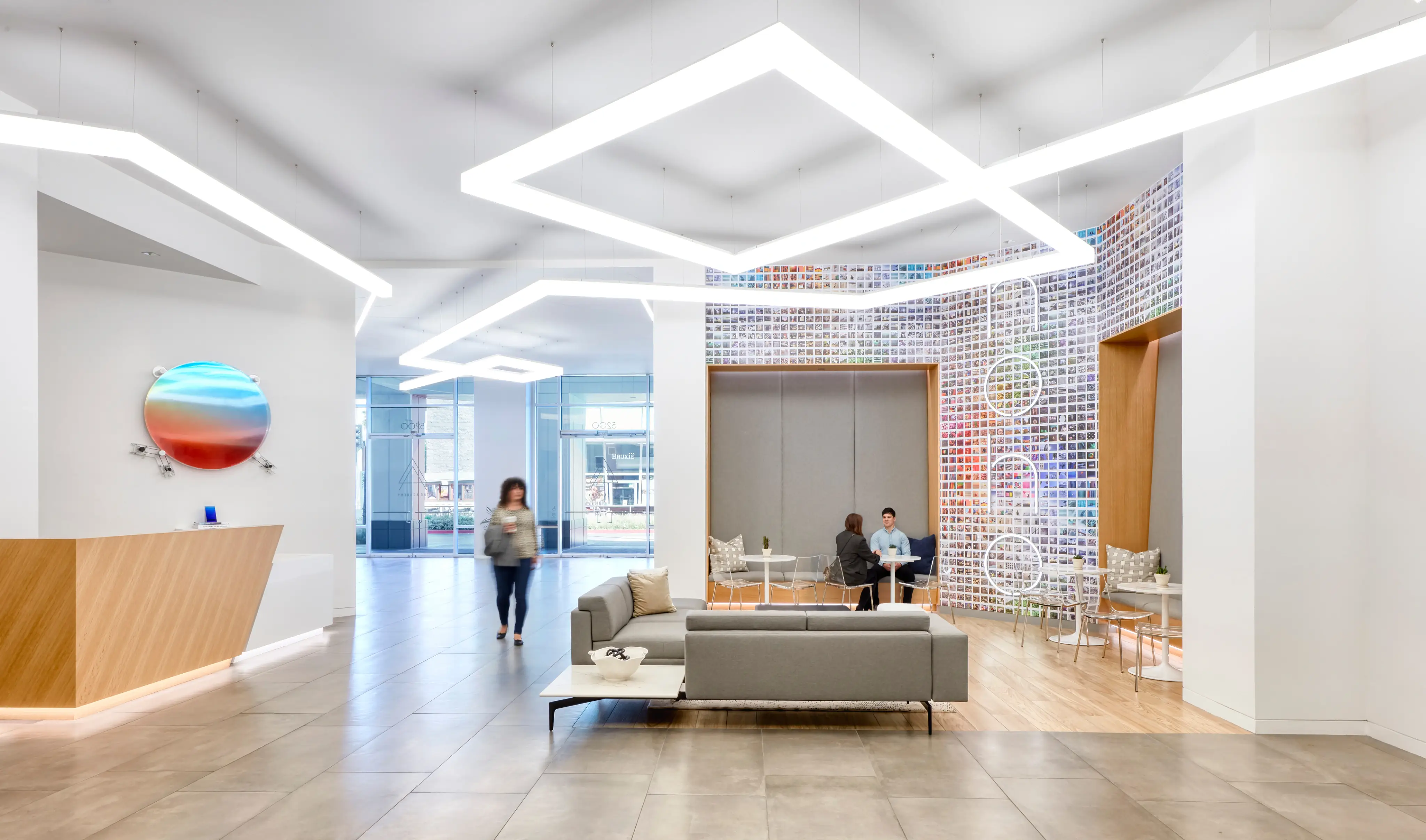 Modern office lobby with geometric ceiling lights and a colorful mosaic wall. A person walks past a seating area where two people converse. Bright and minimalistic.