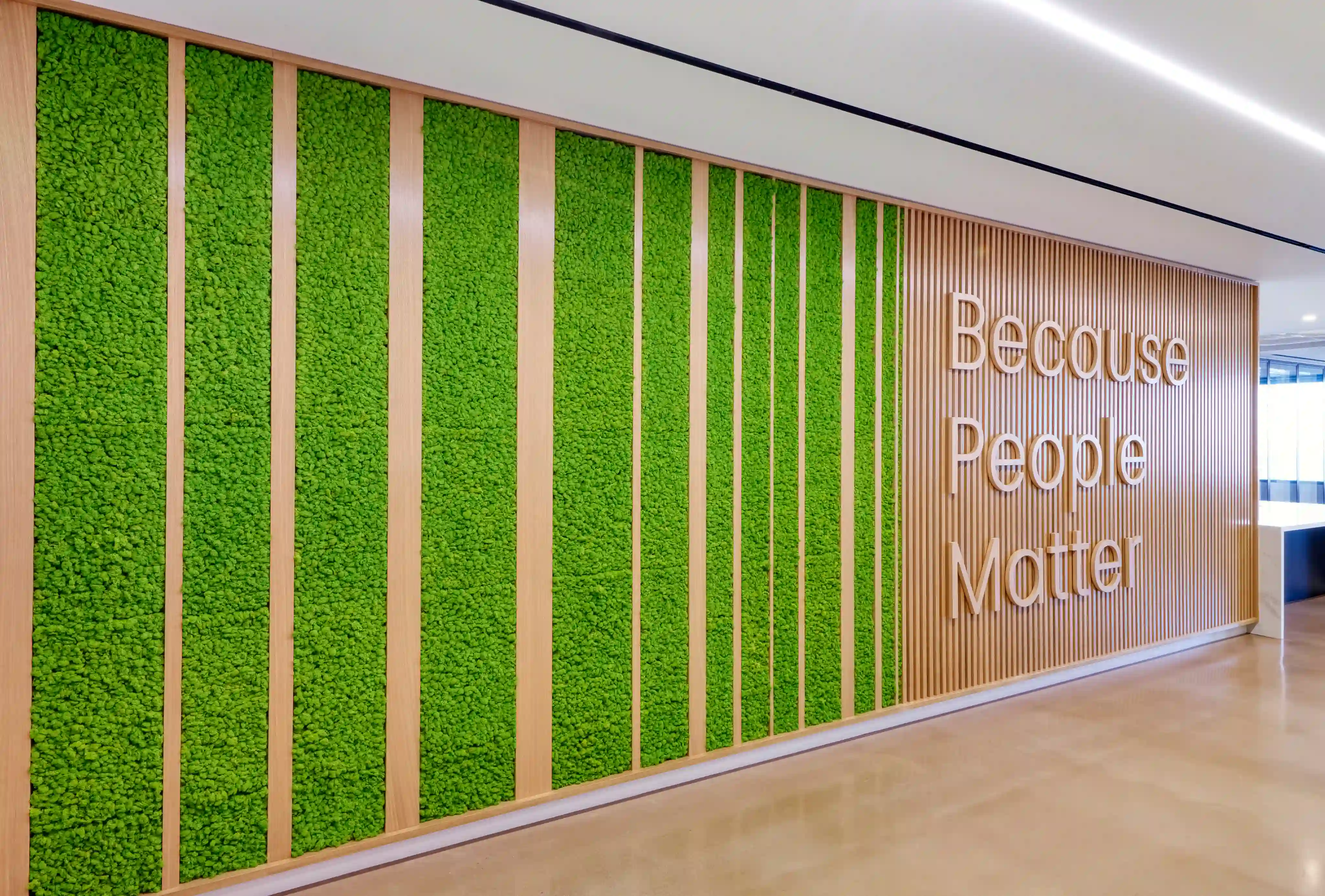 Wall with green moss panels and wooden slats, featuring the words "Because People Matter" in large, white, 3D letters. Bright, modern, and welcoming space.