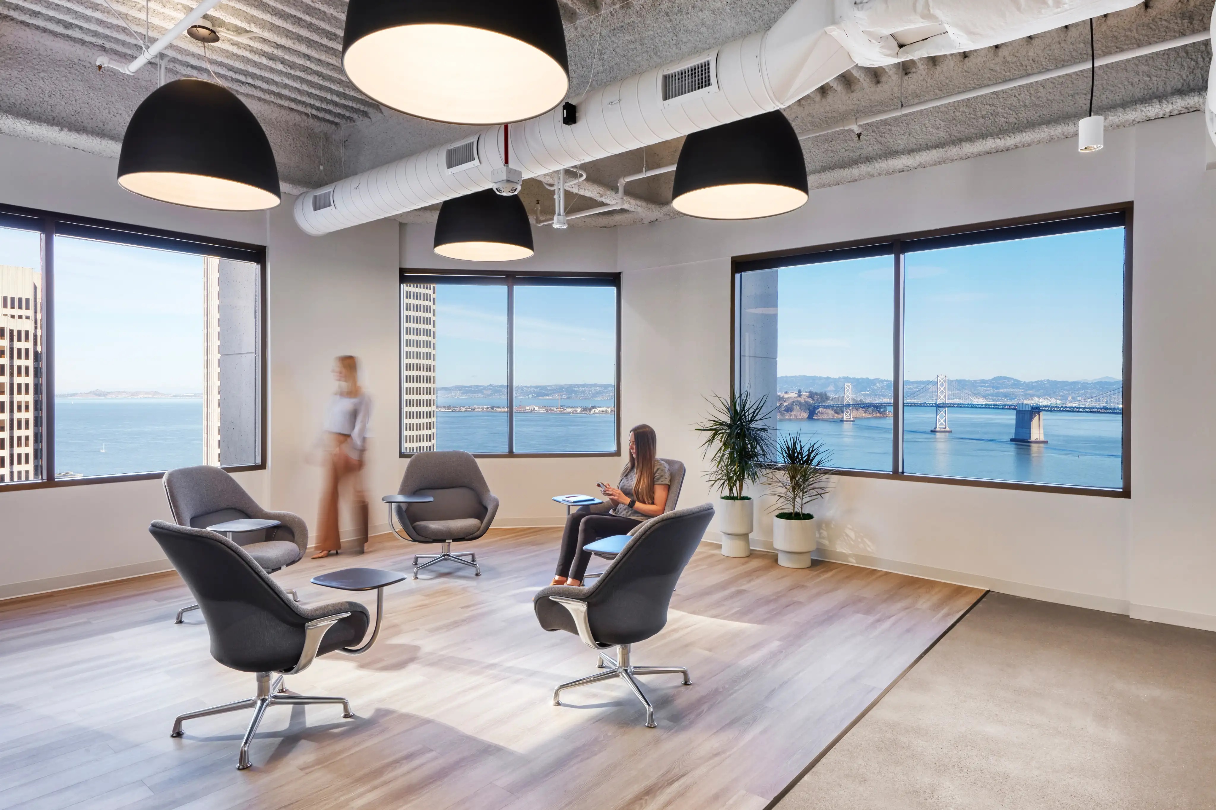 Modern office lounge with large windows showcasing a bay and bridge view. Includes chic seating, two people, potted plants, and sleek lighting. Calm ambiance.